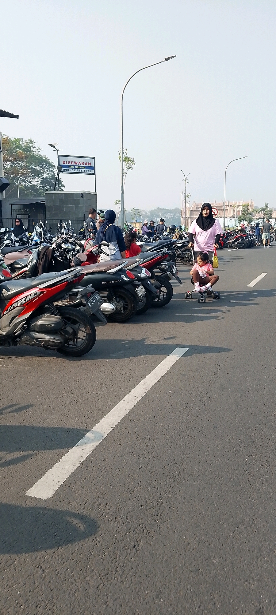 In Gunung Putri, Indonesia, an outdoor scene shows a paved area with numerous parked motorcycles. A woman wearing a pink hijab and a light pink shirt pushes a young child on a small tricycle-like toy. The child is wearing a pink top and brown bottoms. Behind them, other people are visible, some near motorcycles and others in the background, along with trees and a building with a "DISEWAKAN" (rented) sign. The sky is bright and clear, suggesting daytime. The ground is asphalt, marked with a white line.