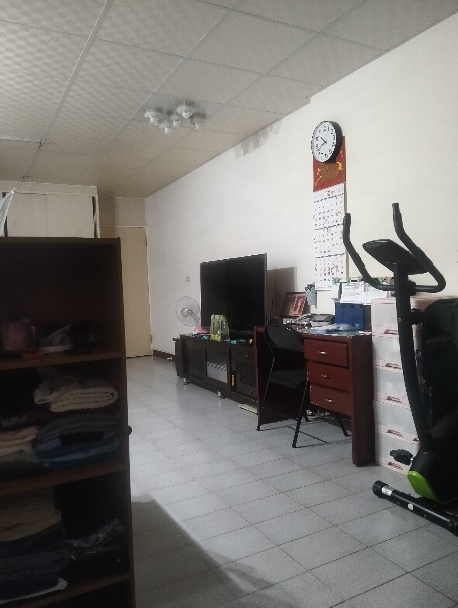 The image shows an indoor scene, likely a living room or a study, featuring a variety of household objects. On the left, a dark brown bookshelf is partially visible, filled with folded clothes and other items. In the center, a black television sits atop a dark wood media console. To the right of the television, a small, silver-colored fan is placed on a shelf. Further right, a wooden desk with red drawers is positioned against the wall. A black folding chair is tucked under the desk. On the wall above the desk, a round clock with black numbers and hands is mounted, accompanied by a calendar for October. A black exercise bike is positioned in the foreground on the right side of the image. The floor is tiled with light gray square tiles. The ceiling is a white, textured paneling system with several light fixtures. The scene appears to be in a residential setting, possibly in Kaohsiung, Taiwan, given the typical interior design elements and the presence of a calendar that may indicate local holidays or customs. There are no people visible in the image.
