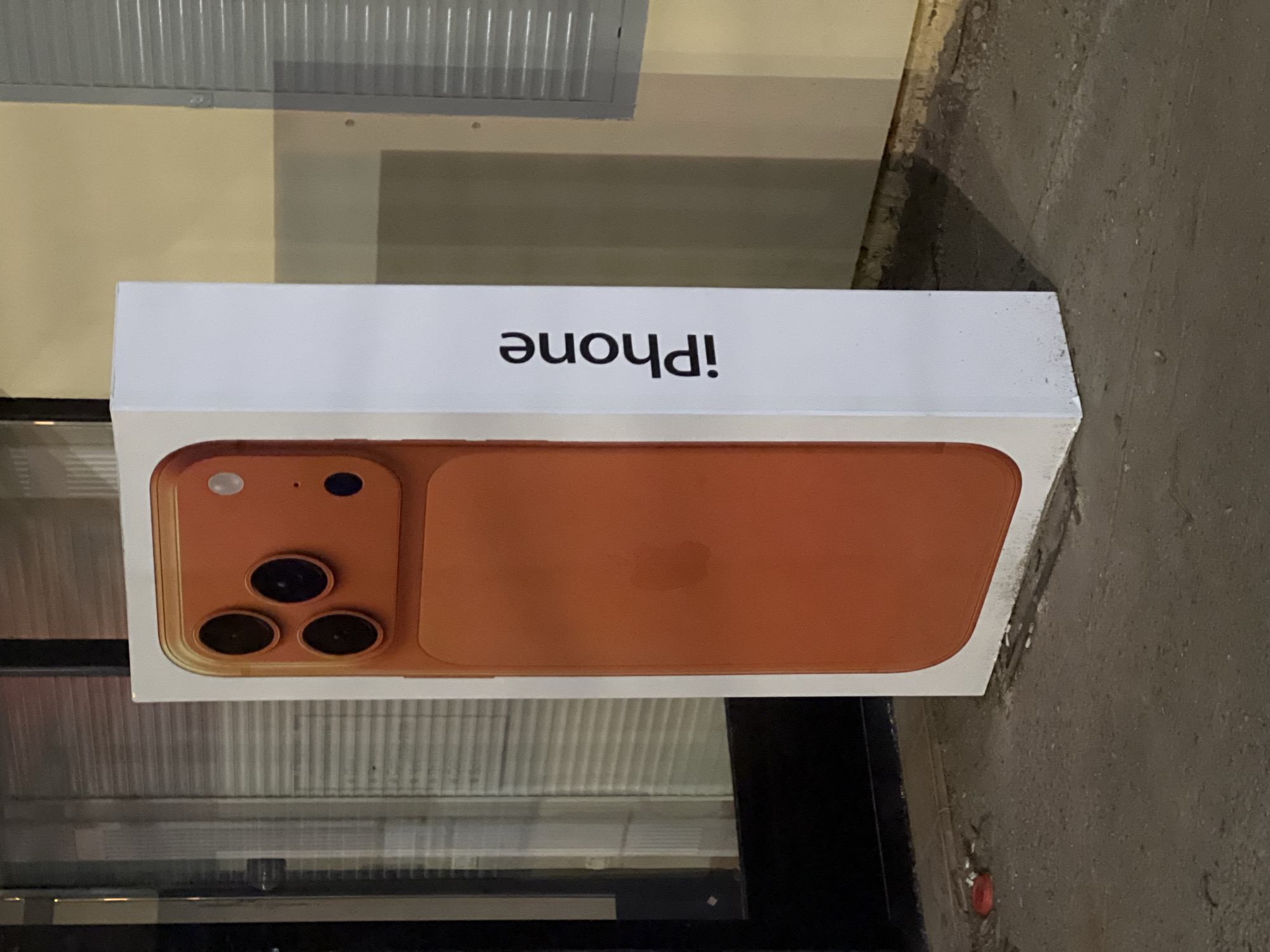 A white iPhone box is centrally featured in this image, appearing to be rotated 90 degrees counter-clockwise. The box rests on a rough, grey concrete or paved surface. An image of an orange or copper-colored iPhone, equipped with a triple-lens camera system on its back, is prominently displayed on the box. The brand name "iPhone" is printed in black text on the side of the box, appearing upside down in the current image orientation.
In the background, above the box, there are horizontal ribbed metal panels, resembling roller shutters or industrial blinds, situated over a pale yellow-beige wall. To the right of the box, the concrete surface extends further, showing textural variations and some light debris. The environment appears to be artificially lit, with reflections visible on the metallic surfaces. No individuals are present, and no particular activity is taking place. The image captures a still life of the product box in what seems to be an urban or industrial outdoor setting in Izhevsk, Russia.