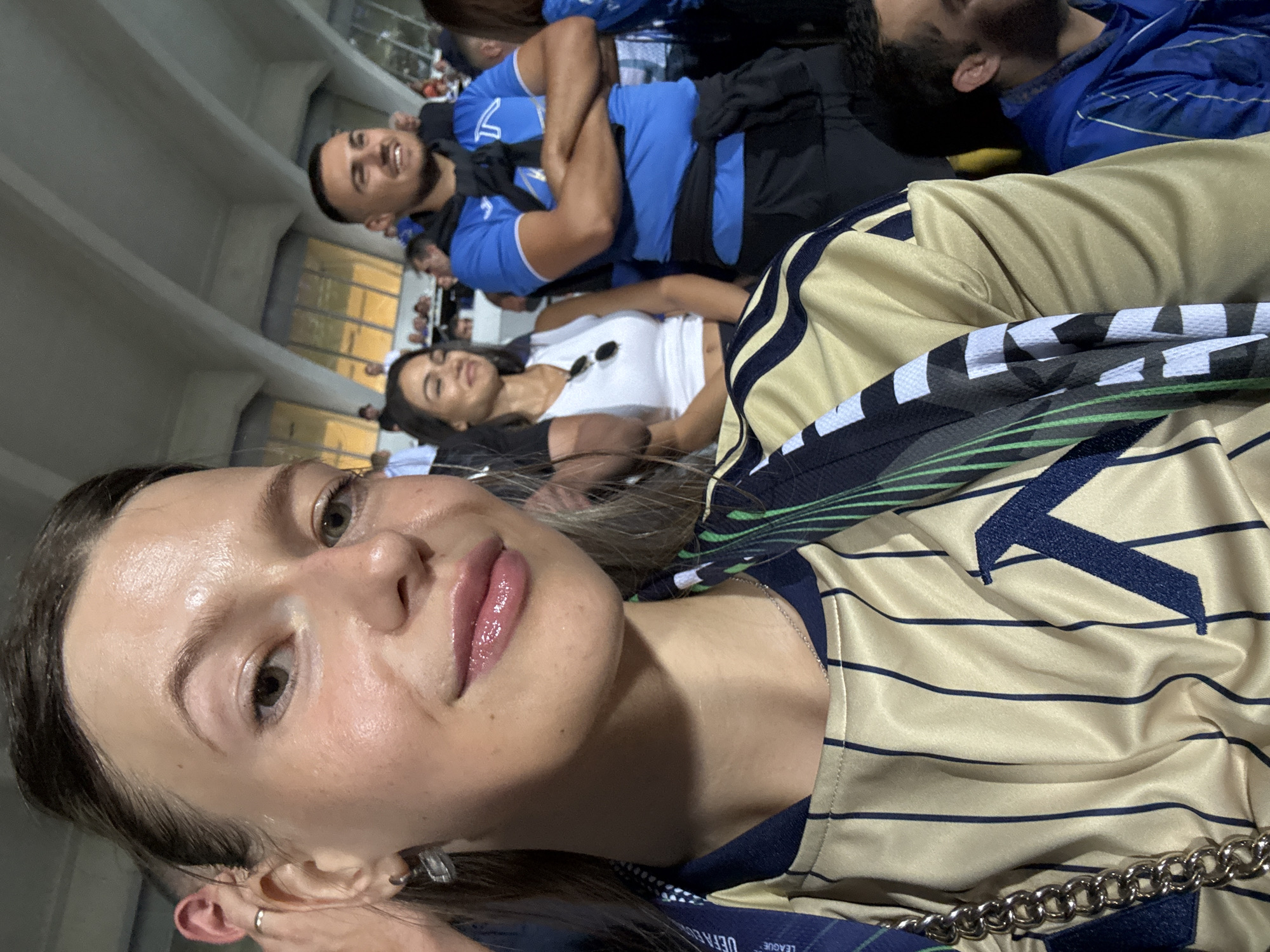 A young woman with blonde hair pulled back and wearing a gold and navy striped jersey is taking a selfie in a stadium. She is smiling and looking at the camera. Her lips are glossy and pink. She is wearing a silver chain necklace and a silver earring in her left ear. A lanyard with "UEFA LEAGUE" printed on it is visible around her neck. Behind her, a man in a blue t-shirt with a navy collar is visible, with his arms crossed. To his left, a woman in a white tank top and sunglasses is also looking to the side. Other spectators are seated in the background, and the stadium's ceiling structure is visible. The overall environment suggests a sporting event.