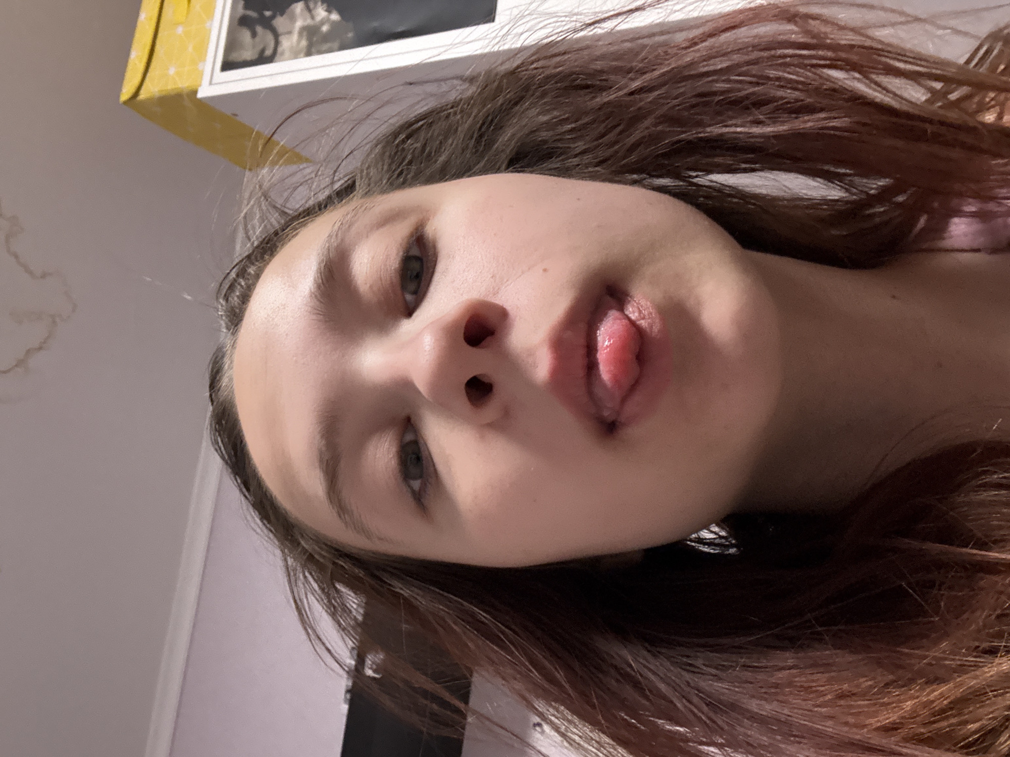 A close-up, angled shot shows a young person with light skin, brown hair, and green eyes. Their tongue is sticking out. The background appears to be an interior space with a white wall and a cabinet. The photograph was taken at an angle, obscuring the details of the location in Прикамский, Russia. No interactions are visible.