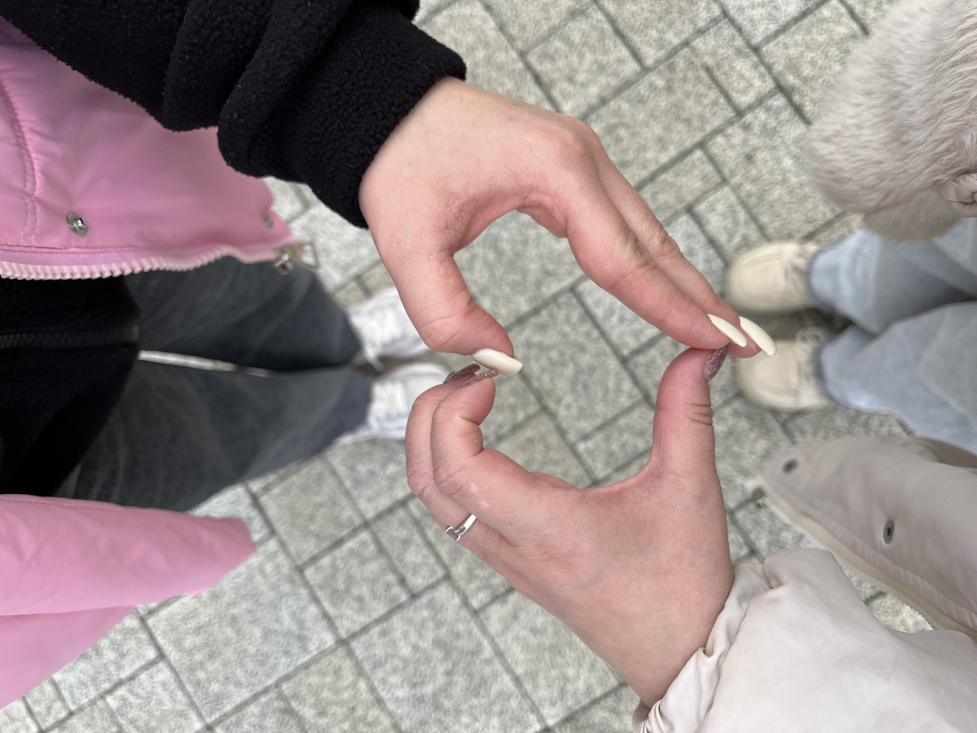 Two people stand on a paved surface, likely a sidewalk or plaza, creating a heart shape with their hands. The person on the left wears a black sweater, a pink vest, and grey jeans with white sneakers. The person on the right wears a white jacket and light blue jeans with light-colored sneakers. A small, white object is held between the fingertips of the two people. The ring finger of the right hand bears a silver ring. The scene is composed from a top-down perspective. The location is Novopokrovskaya, Russia.