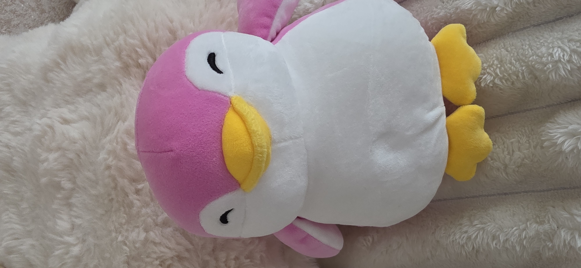 A plush toy penguin with pink and white coloring is positioned on a textured, fuzzy, cream-colored surface. The penguin has a pink head and back, with a white belly and chest. Its beak is bright yellow, and it has black eyes. Its feet are also yellow. The surface beneath the penguin appears to be a soft blanket or rug. There is no visible indication of the city of Sumgayit, Azerbaijan, in the image.