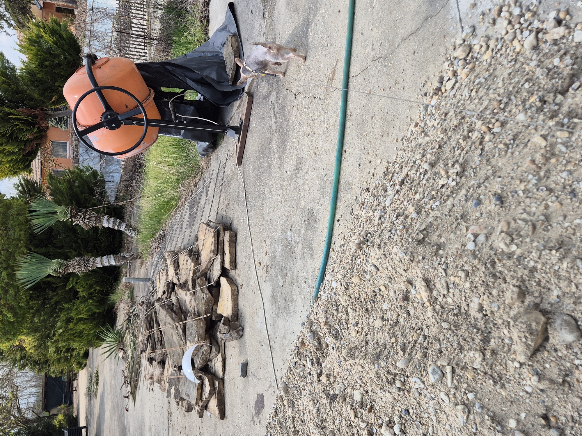 The image shows an outdoor scene in Pavlikeni, Bulgaria. The setting is a concrete-paved area. An orange concrete mixer with a black wheel sits near a pile of stacked stones and a pile of sand. A small dog stands near the mixer. A green hose runs across the concrete. There are bushes and trees in the background. A partially visible building can also be seen in the background.