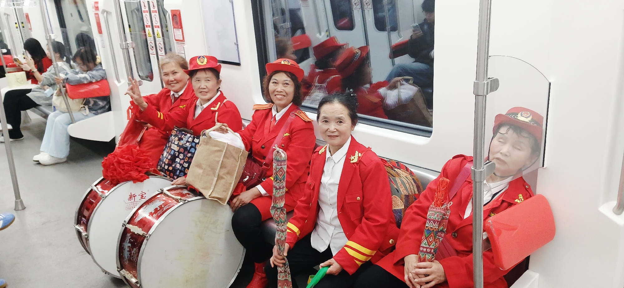 The image shows a group of five older women in matching red uniforms and hats, seated on a subway train. The women are positioned along one side of the train car, with two large drums beside them. Other passengers are visible in the background and through the train window. Based on the drum labels and other visual clues, the location is likely Shapingba District, China. One of the women at the end of the row is asleep.