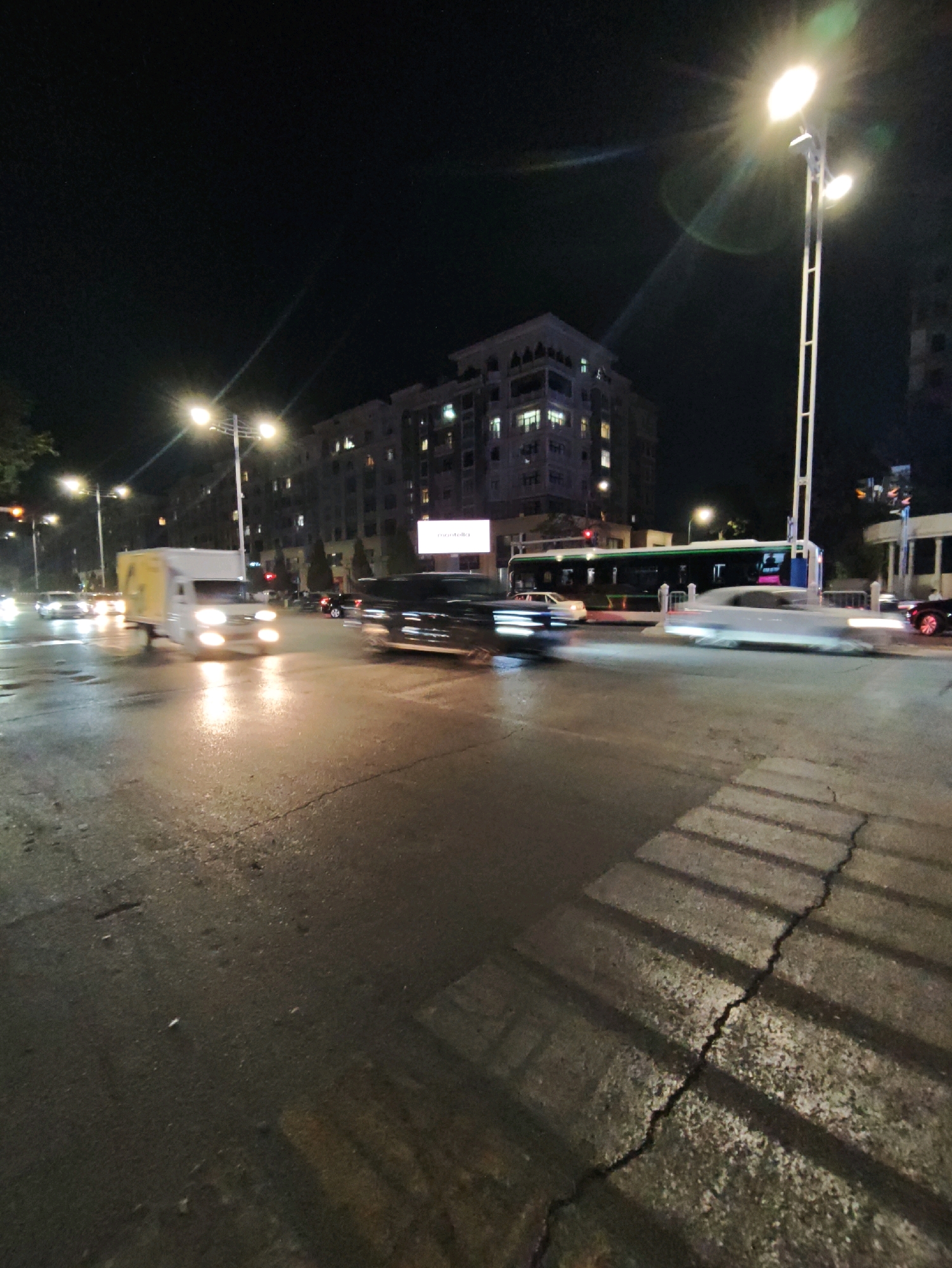 This is a night-time, street-level, wide-angle shot of a busy intersection in Tashkent, Uzbekistan. The street is wet and reflects the bright streetlights and vehicle headlights. In the foreground, a cracked asphalt road is visible, with a prominent shadow of a fence cast across it. Several vehicles are in motion, indicated by motion blur. A white truck with yellow markings is visible on the left side of the image, with its headlights illuminated. Further down the road, a bus with green and white livery is seen, along with other cars. In the background, multi-story buildings are illuminated with interior lights. A tall street lamp with multiple bulbs stands prominently on the right side of the image, emitting a bright glare. The overall impression is of a vibrant urban environment at night.