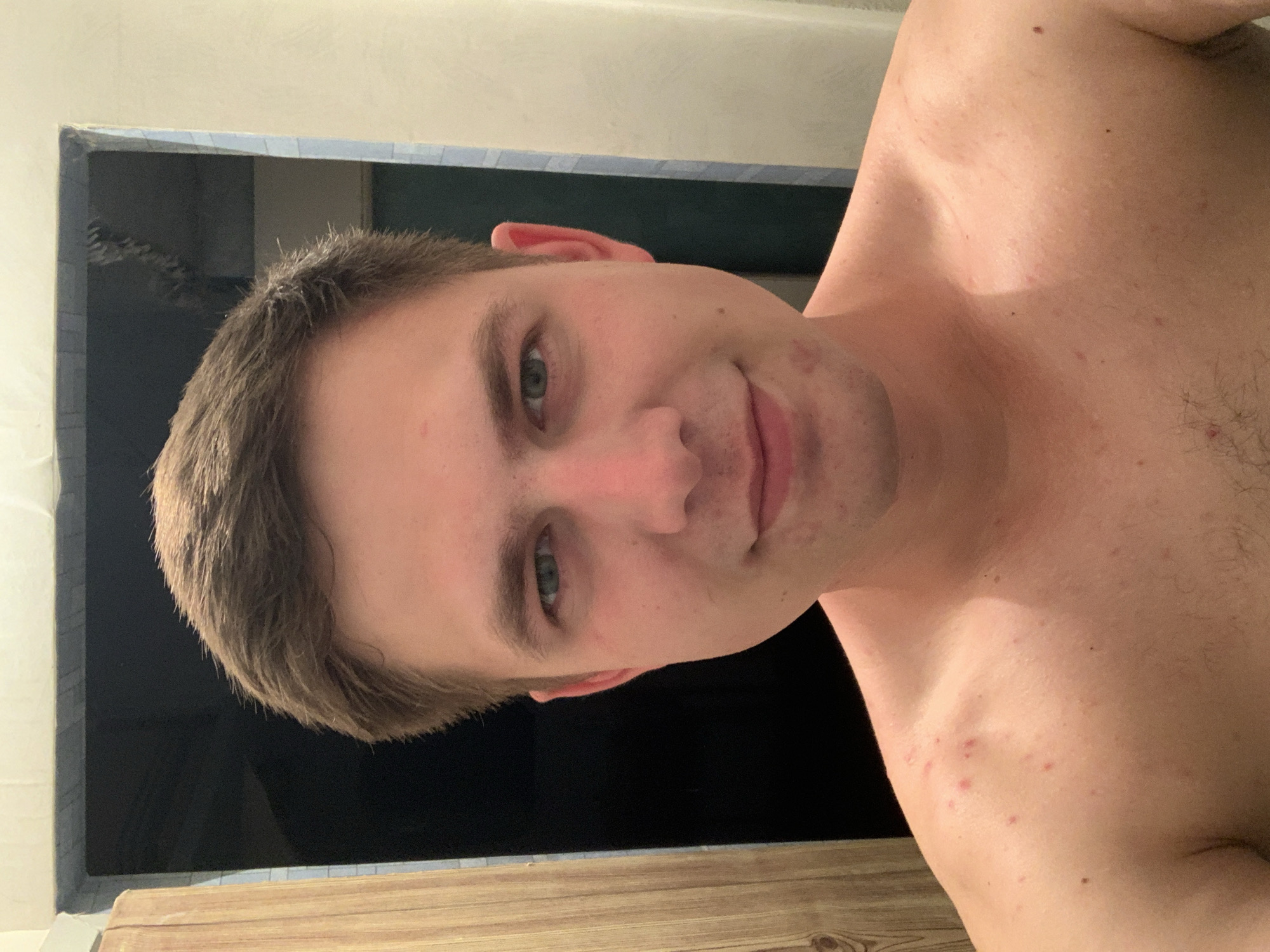 An image shows a young Caucasian male with short brown hair and green eyes. He is looking at the camera and smiling slightly. The man is shirtless and appears to have some acne on his shoulders and chest. The background is blurry and appears to be a bathroom with a wooden door on the left and a tiled wall on the right. The lighting is dim, casting a shadow on the man's face.