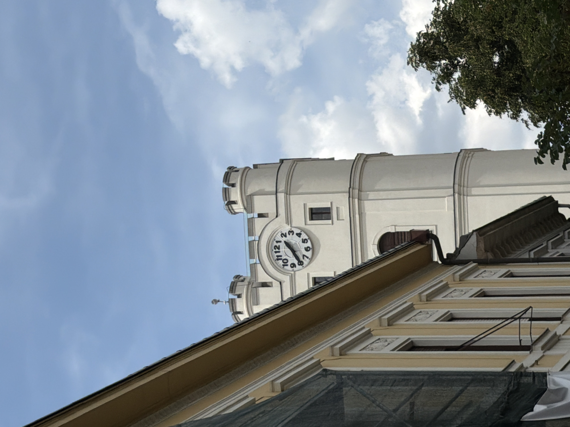 The image shows a view of the Reformed Great Church of Debrecen, Hungary, from a low angle looking upwards. The church features a prominent clock tower with a white facade and a circular clock face. The clock shows the time as approximately 9:55. In the foreground, a yellow building with ornate window frames is visible, partially obscuring the view of the church. The sky is blue with scattered white clouds, and the top of a green tree can be seen on the right side of the image. The scene appears to be taken on a bright, partly cloudy day.