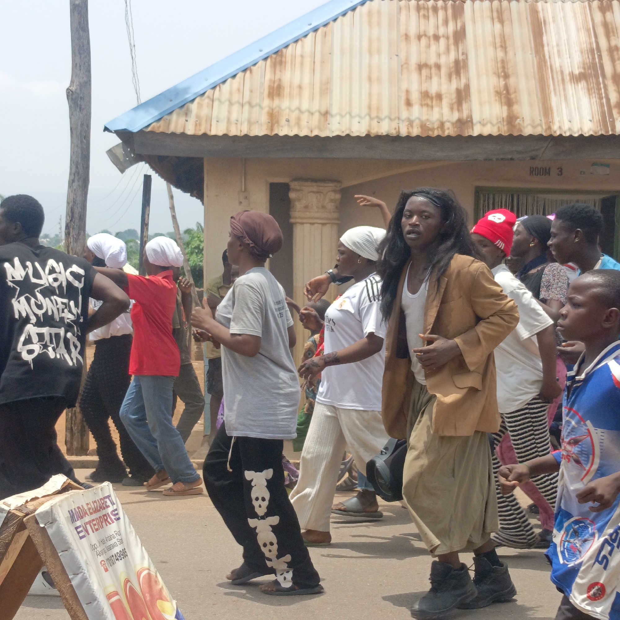 A group of people are walking down a street. A man with long hair wears a tan jacket and light-colored pants, walking with the group. A sign advertising "MADA ELIZABETI ENTERPRISE" is visible on the left. A building with a corrugated metal roof is in the background with a sign that reads "ROOM 3". The sign also contains the text "Opp. Jilbab Andaha Ror Akwanga Nasarawa State", identifying the location as Akwanga, Nasarawa State, Nigeria.