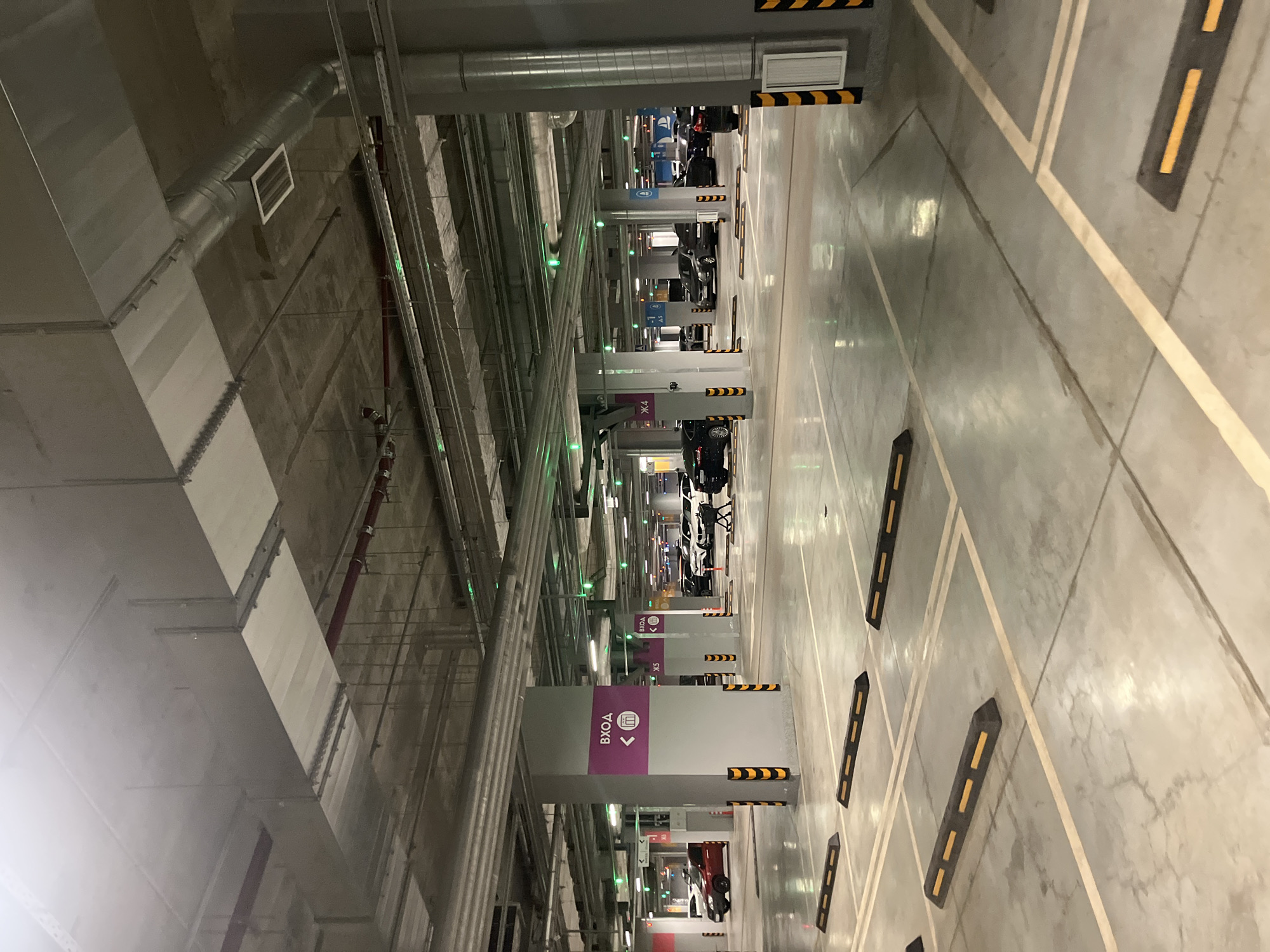 The image captures an indoor, multi-level parking garage located in Saint Petersburg, Russia. No individuals are present in the scene.
The garage features numerous grey concrete pillars, supporting an exposed concrete ceiling. Several pillars display prominent purple signs with white Cyrillic text "ВХОД" (Entrance), an arrow pointing left, and a building icon. Other pillars are marked with alphanumeric identifiers such as "Ж4" and "Ж5". The lower sections of the pillars are protected by black and yellow striped bumper guards.
The floor consists of polished concrete, exhibiting yellow painted lines that delineate parking spaces and driving lanes. Black and yellow rubber parking stops are positioned at the end of each parking bay. The concrete floor shows signs of wear, including visible cracks and minor fluid streaks. Overhead, the industrial ceiling infrastructure is exposed, comprising large rectangular silver ventilation ducts, smaller circular pipes, red fire suppression piping, and multiple white fluorescent light fixtures. Numerous small green LED lights, consistent with parking space occupancy indicators, are embedded in the ceiling structure.
Approximately 10 to 15 vehicles, primarily SUVs and sedans of various makes and colors including red, black, white, and grey, are parked in the mid-ground and background. A single shopping cart is visible near a black parked vehicle in the mid-right portion of the image. The environment is uniformly illuminated by artificial light sources, which cast reflections on the polished floor surface.