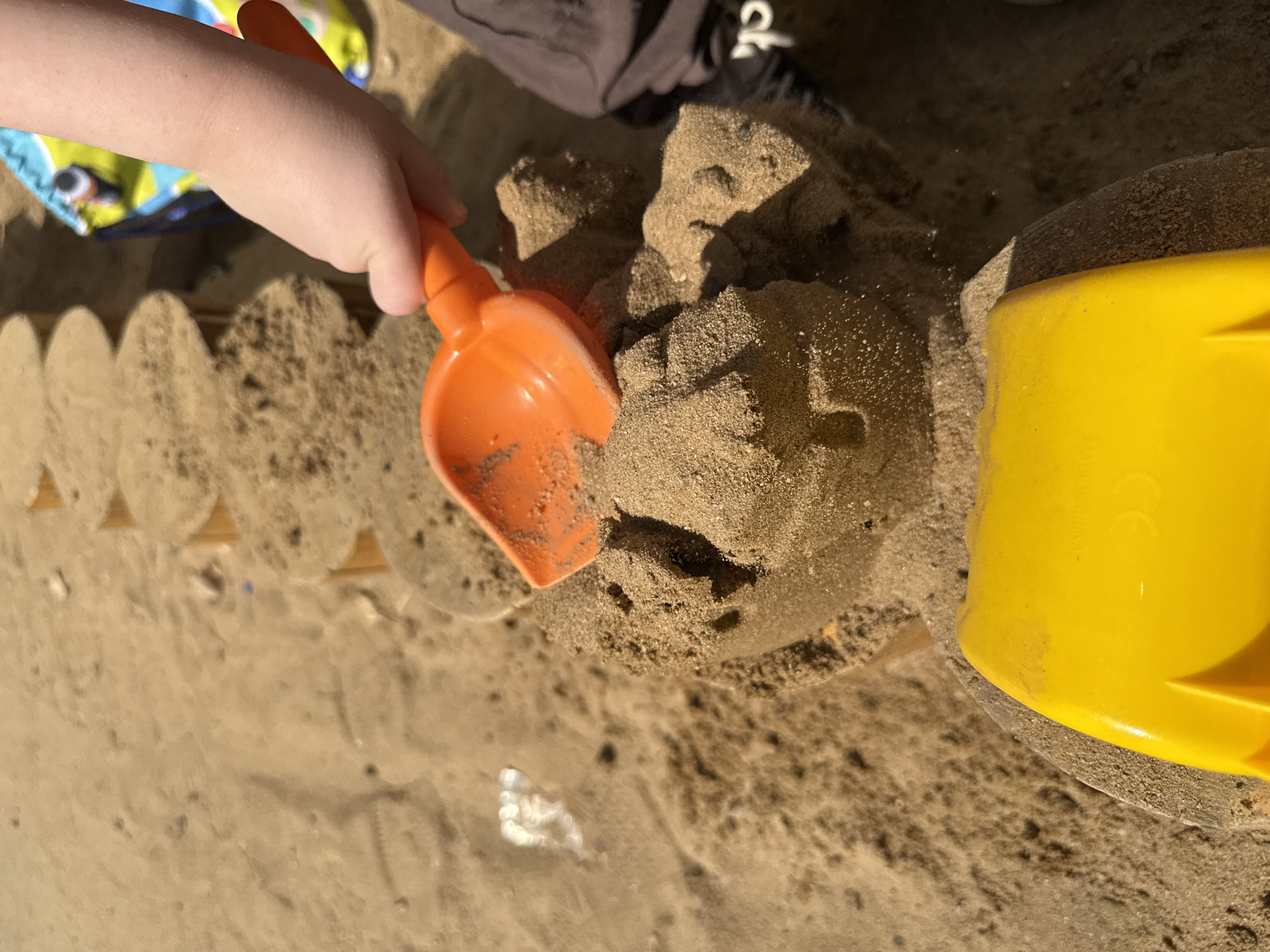 A child's hand holds an orange plastic shovel to manipulate a sandcastle in a sandbox. A yellow plastic bucket sits to the right. The sandbox has a scalloped wooden border. Part of a garment with a colorful pattern is visible on the left and dark trousers on the upper center. The scene is in a sandbox with tan sand. The location is unknown.
