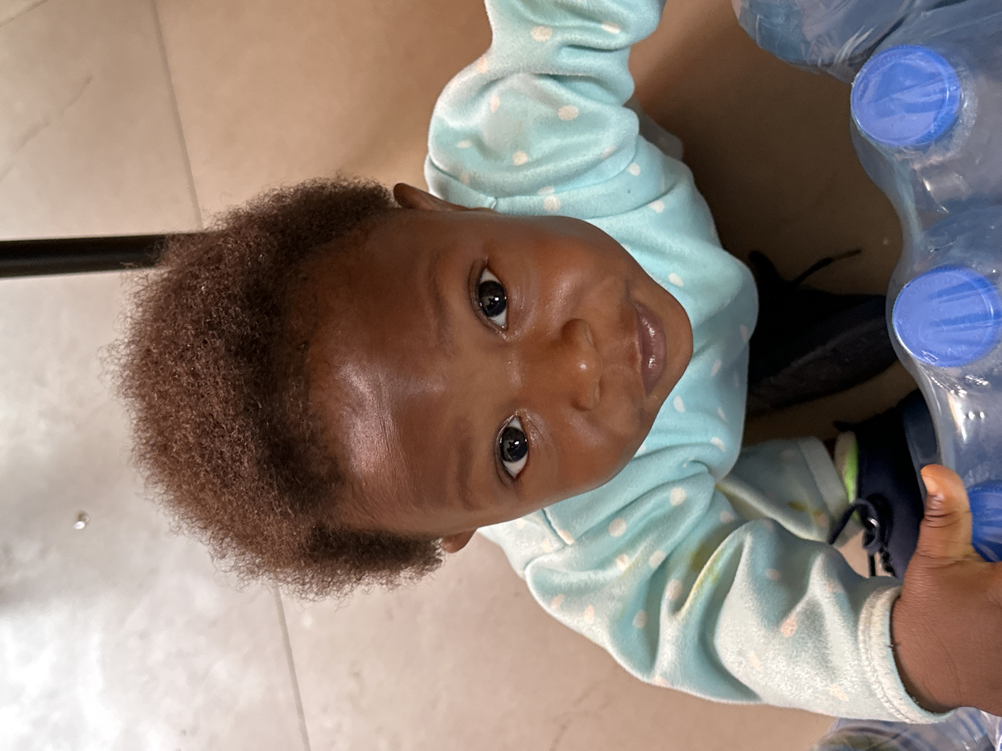 This vertical image, captured in Jos, Nigeria, features an infant or toddler looking directly upwards at the camera. The child has dark skin and short, dark, curly hair, with wide, dark eyes expressing curiosity or a slight smile. They are wearing a light blue long-sleeved top patterned with white polka dots.
The setting is indoors, with a light-colored, possibly beige, tiled floor forming the background. On the right side of the frame, a package of multiple plastic water bottles with blue caps, wrapped in clear plastic, is visible, which the child appears to be reaching towards or interacting with. A dark object, likely a shoe or part of an adult's foot, is partially seen near the child's right arm and the water bottles. A thin black vertical element, possibly a furniture leg or railing, is present on the left. The lighting suggests an indoor, well-lit environment. No discernible text is visible on any of the subjects or objects. The photograph captures a candid moment of the child's engagement with their immediate surroundings.