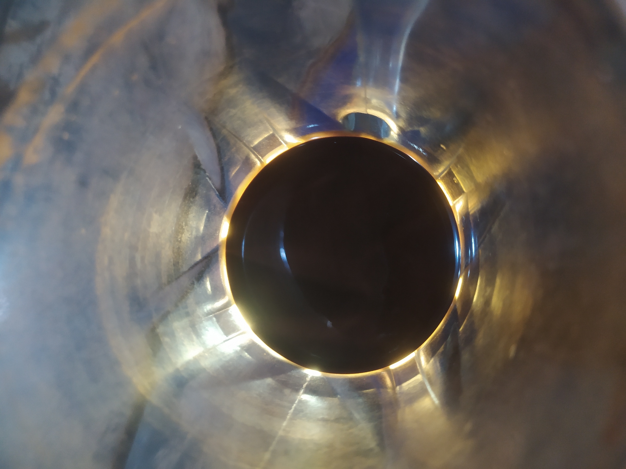 The image shows a close-up, interior view of a metallic pipe or cylindrical object. The surface appears to be made of polished metal, reflecting light and creating a sense of depth and texture. The lighting highlights the curved surfaces, with bright yellow and white highlights juxtaposed against darker, shadowed areas. In the center of the image, a dark, circular void is visible, suggesting the opening of the pipe or an object within it. The overall impression is abstract and focused on the interplay of light and form within a confined space. There is no indication of a specific location in Tuntang, Indonesia, nor any people, actions, or interactions depicted.