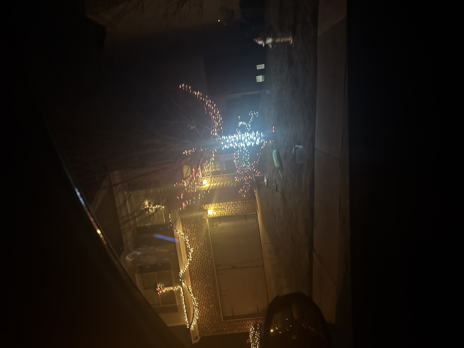 A nighttime view from inside a vehicle captures a brick building adorned with holiday lights. Garlands and strings of lights outline windows and architectural features. A decorated tree stands prominently, its branches illuminated by a dense cluster of white and red lights. A fire hydrant is visible in the background. The darkness of the vehicle's interior frames the festive scene.