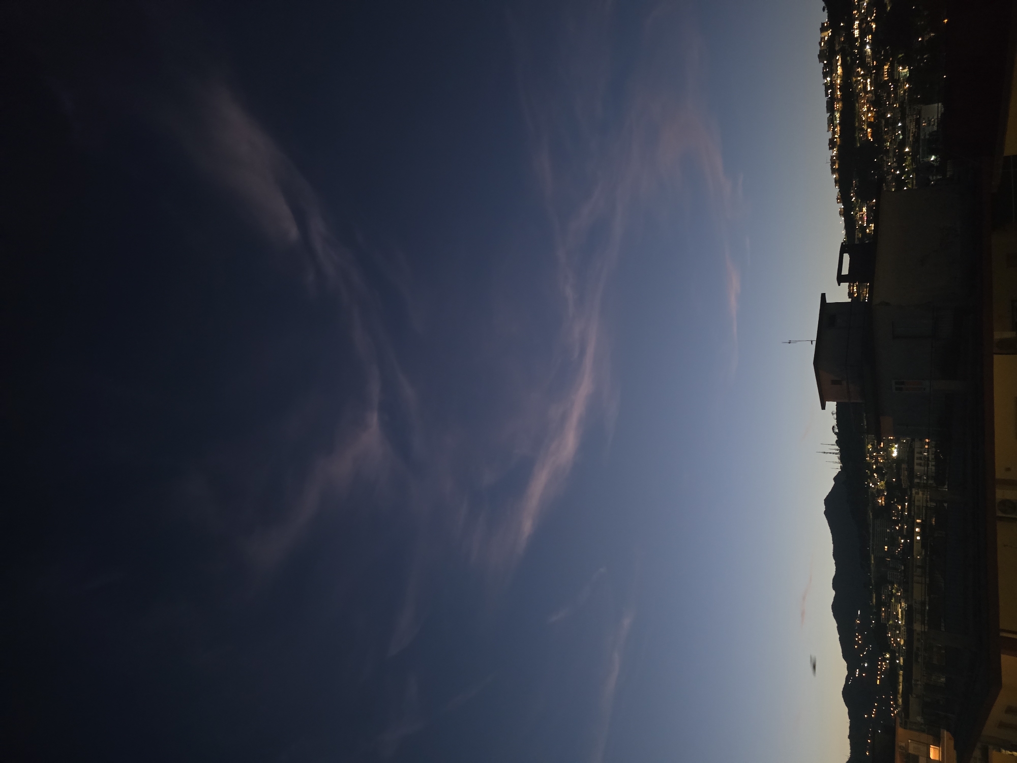 The image depicts an urban landscape at twilight, characterized by a gradient sky and illuminated city lights against a mountainous backdrop. The upper portion of the sky is a deep indigo, transitioning to a lighter blue and subtle orange-pink hues closer to the horizon. Wispy, elongated clouds, faintly illuminated with pink and white, are present across the middle and upper sections of the sky.
In the midground, a distinct silhouette of irregular mountain peaks forms the horizon line. Below this, the city of Salerno, Italy, extends across the slopes and into the foreground. Numerous artificial lights, predominantly yellow and orange, are visible across the city, outlining buildings and roads on the dark hillsides.
The immediate foreground features dark silhouettes of building rooftops. A prominent, multi-story building with a flat roof and a smaller, box-like structure on top is visible in the lower-middle right, along with another rectangular structure resembling a chimney or vent. No individuals or specific activities are discernible within the frame. The overall lighting is dim, relying on ambient twilight and the city's artificial illumination.