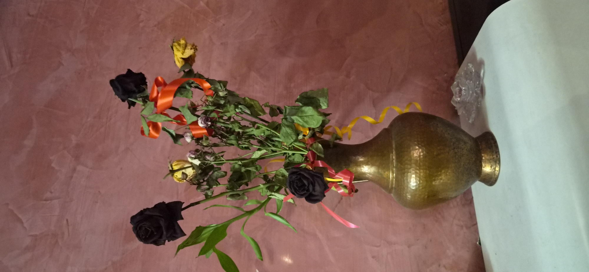 The image displays a still life composition featuring a bouquet of roses in a decorative vase, set against a textured wall. The main subject is a rustic metal vase, likely hammered brass or copper, with a wide body and a narrow neck. It rests on a flat, white surface, possibly a table or shelf.
Within the vase, a bouquet of roses is arranged with green foliage. Notable among the flowers are several black roses, some faded yellow roses that appear withered, and other smaller, pale blossoms that also show signs of wilting. Bright orange and yellow ribbons are intertwined among the roses and stems, providing a vibrant contrast to the somber tones of some of the flowers and their decaying state. One prominent red-orange ribbon curls down the left side of the arrangement, while thinner yellow ribbons spiral around the top of the vase.
The background consists of a textured reddish-brown wall, possibly finished with stucco or a similar decorative plaster, displaying subtle variations in color and pattern. To the right of the vase, on the white surface, a small, clear, crystalline decorative object, possibly glass or plastic, is visible.
The scene is indoors; no specific activity is taking place as it's a static arrangement. The time of day and weather are not evident. The overall aesthetic combines elements of classic still life with a touch of melancholy due to the withered flowers, brightened by the festive ribbons. No visible text is present.