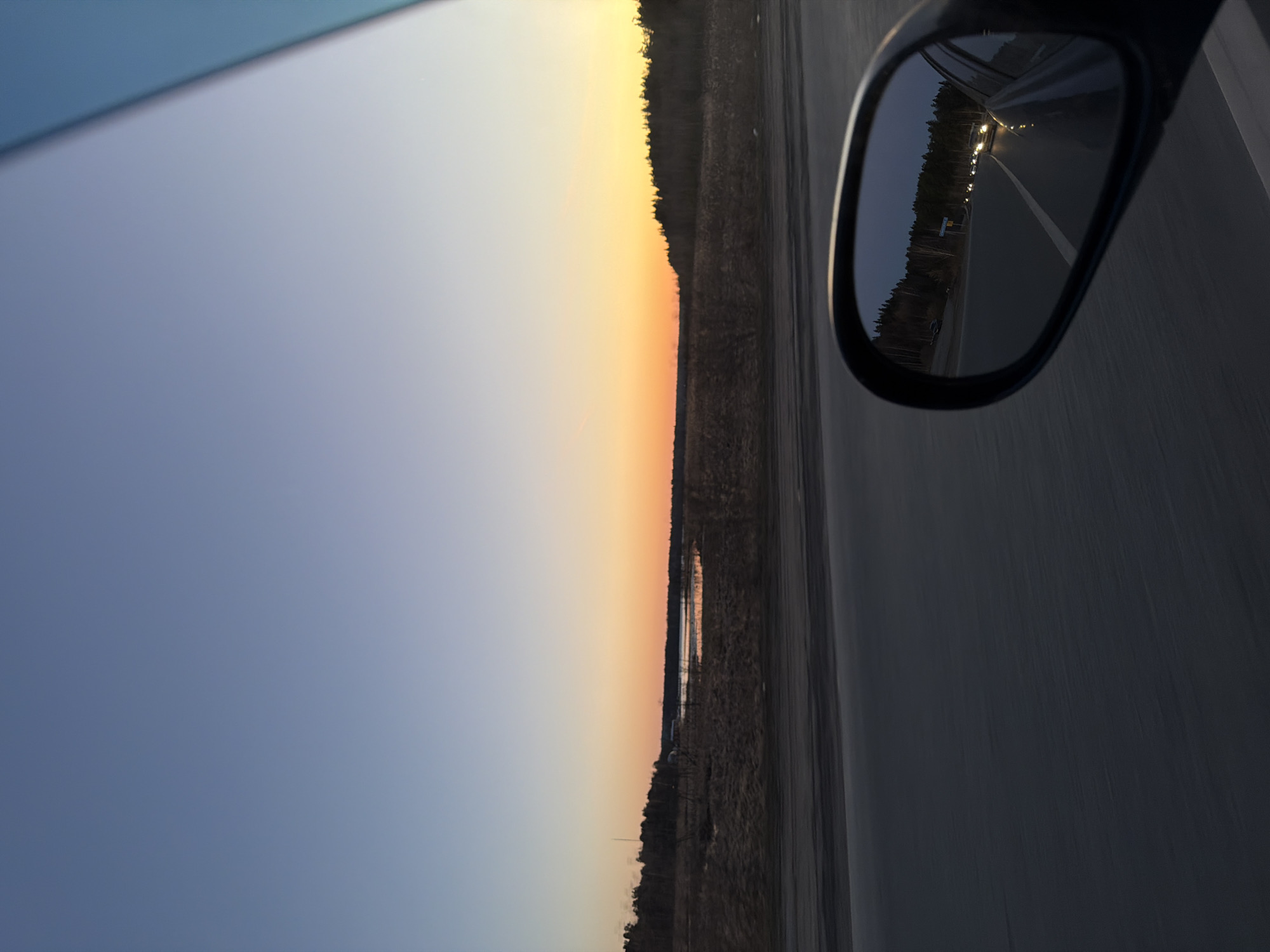 The image captures a sunset view from inside a moving vehicle, likely on a highway near Izhevsk, Russia. The sky transitions from a light blue to orange and yellow hues at the horizon, indicating sunset or sunrise. A treeline is visible at the horizon. The road is visible on the right side of the photo, and a side mirror reflects the road behind the car. The lights of other vehicles are visible in the side mirror. The time of day appears to be dusk.