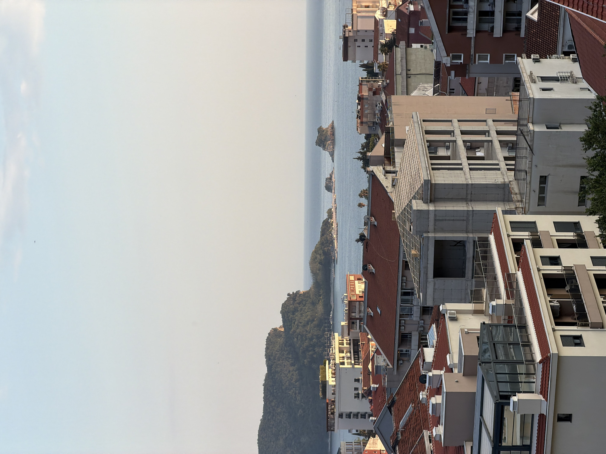 A sideways aerial shot shows the coastal city of Budva, Montenegro. The scene is divided between the sky and the sea on the left side, and closely built houses and other buildings on the right. The sea extends towards the horizon and is populated with small islands. Buildings in the urban landscape have red tile roofs and light-colored walls.