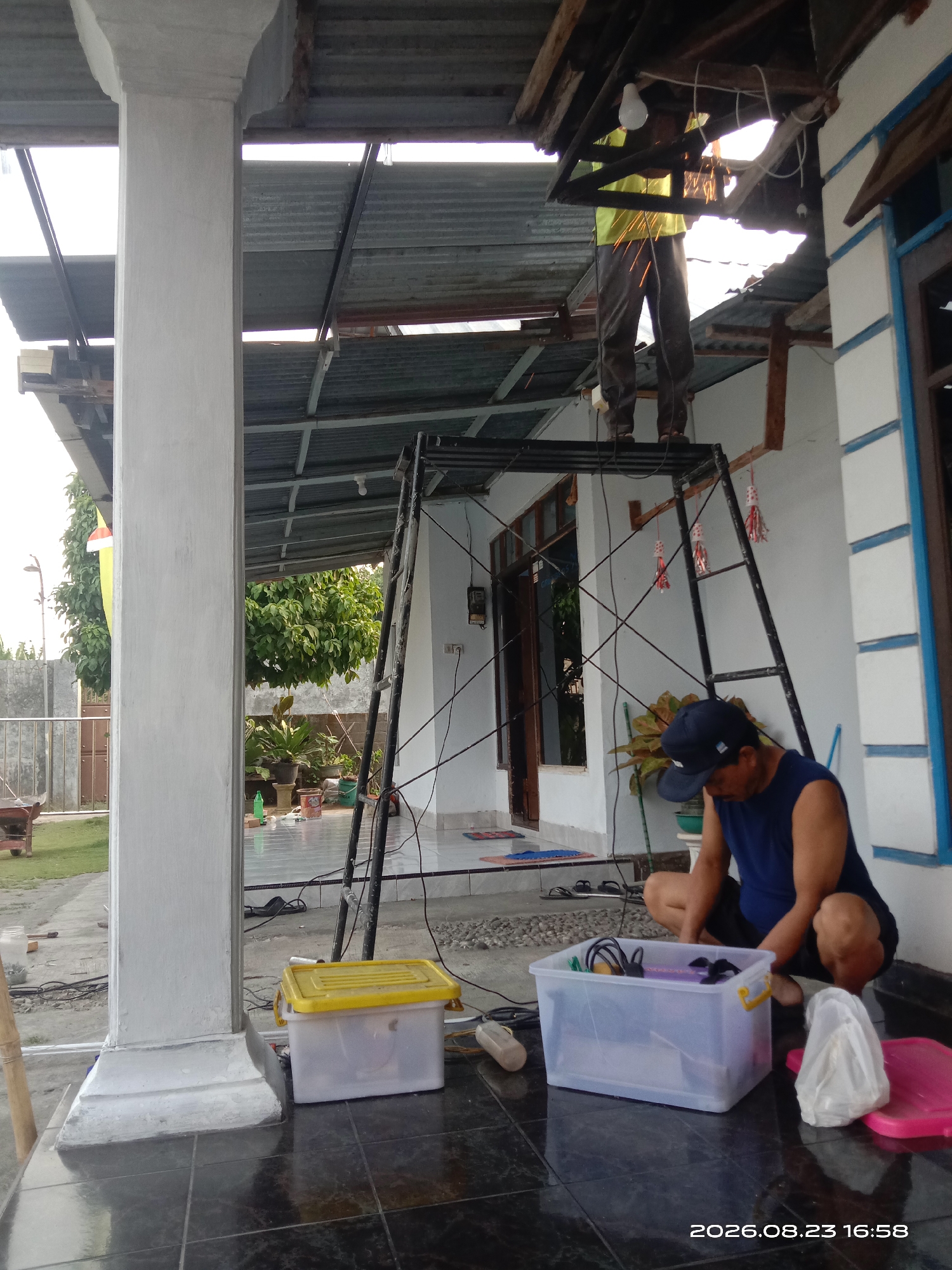 In Banyuarang, Indonesia, a man in a blue tank top and black shorts is crouched on a tiled porch, examining the contents of a translucent plastic bin. The bin, which has yellow handles, contains various items including what appear to be tools or electrical components. Beside it is a pink lid, and further to the left, a yellow-lidded plastic container. The man wears a dark blue baseball cap. In the background, a person in a yellow shirt and dark pants is standing on a metal scaffolding structure, engaged in work that is producing sparks, suggesting metal cutting or grinding. This individual is positioned beneath an exposed, unfinished roof structure made of corrugated metal and wood. Electrical wires are visible, running across the scene. To the left of the frame, a large, light-colored concrete pillar stands prominently, with glimpses of a grassy yard and trees beyond. The overall scene depicts an outdoor construction or repair project. The timestamp on the image indicates August 23, 2026, at 16:58.