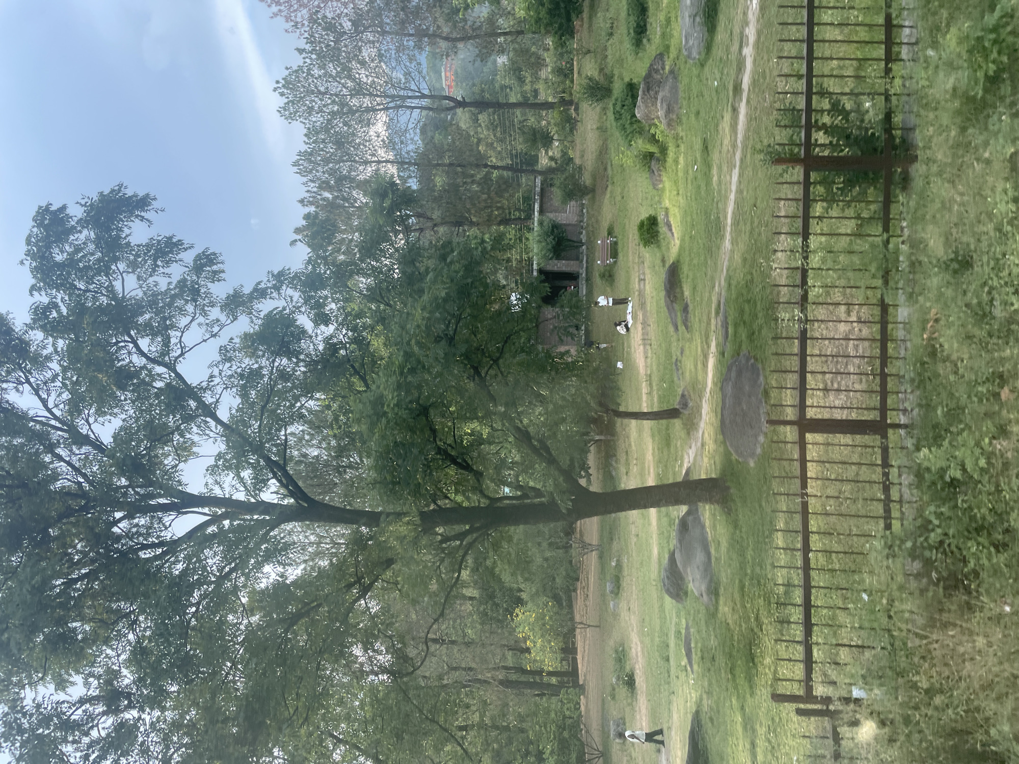 The image depicts a park-like setting with trees, grass, and scattered rocks. A fence is partially visible on the right side of the frame. In the distance, there are buildings and trees suggesting an urban environment. In the middle ground, there is a small structure and two figures wearing white clothing. A person is walking along a path in the foreground. The scene is located in Chetru, India.