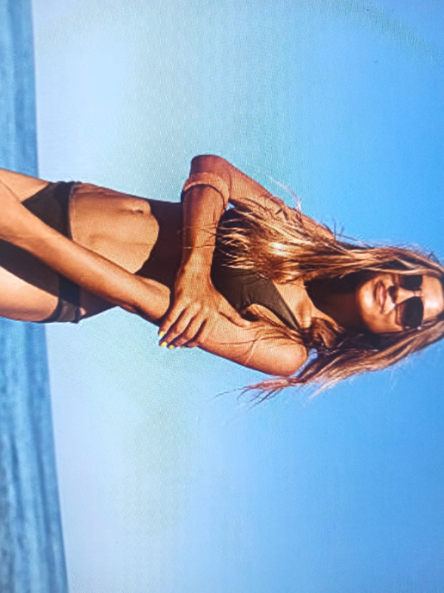 A woman is posing on a beach, leaning back against the sand with the ocean and sky in the background. She is wearing a dark bikini and sunglasses, and her long, blonde hair is flowing around her. Her body is tanned, and she appears to be relaxed and confident. The sun is shining brightly, casting a warm glow on her skin. The scene suggests a leisurely day at the beach, enjoying the warm weather and the ocean breeze. There is no visible text in the image.