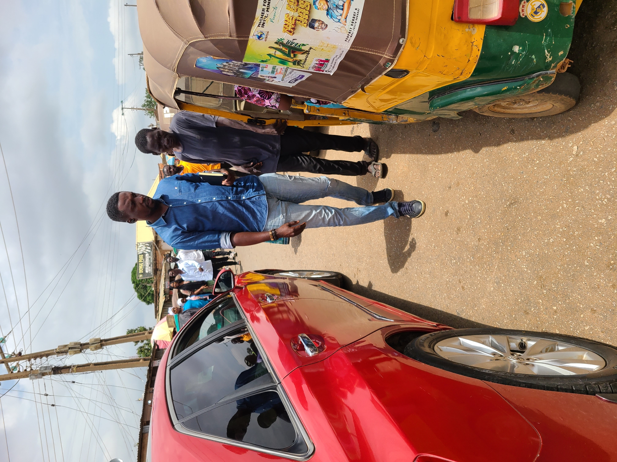 The image shows a man standing on a street next to a red car and a yellow-green auto rickshaw in an urban area. Other people are standing in the background. The auto rickshaw has a poster on its roof for "HIGHER FO INT'L MIND" presents, a "PROPHET SUNDAY & EVELYN VONEBUT" anniversary. There is also a storefront visible in the background with the name "DENGE POSE." Electrical lines are visible above.