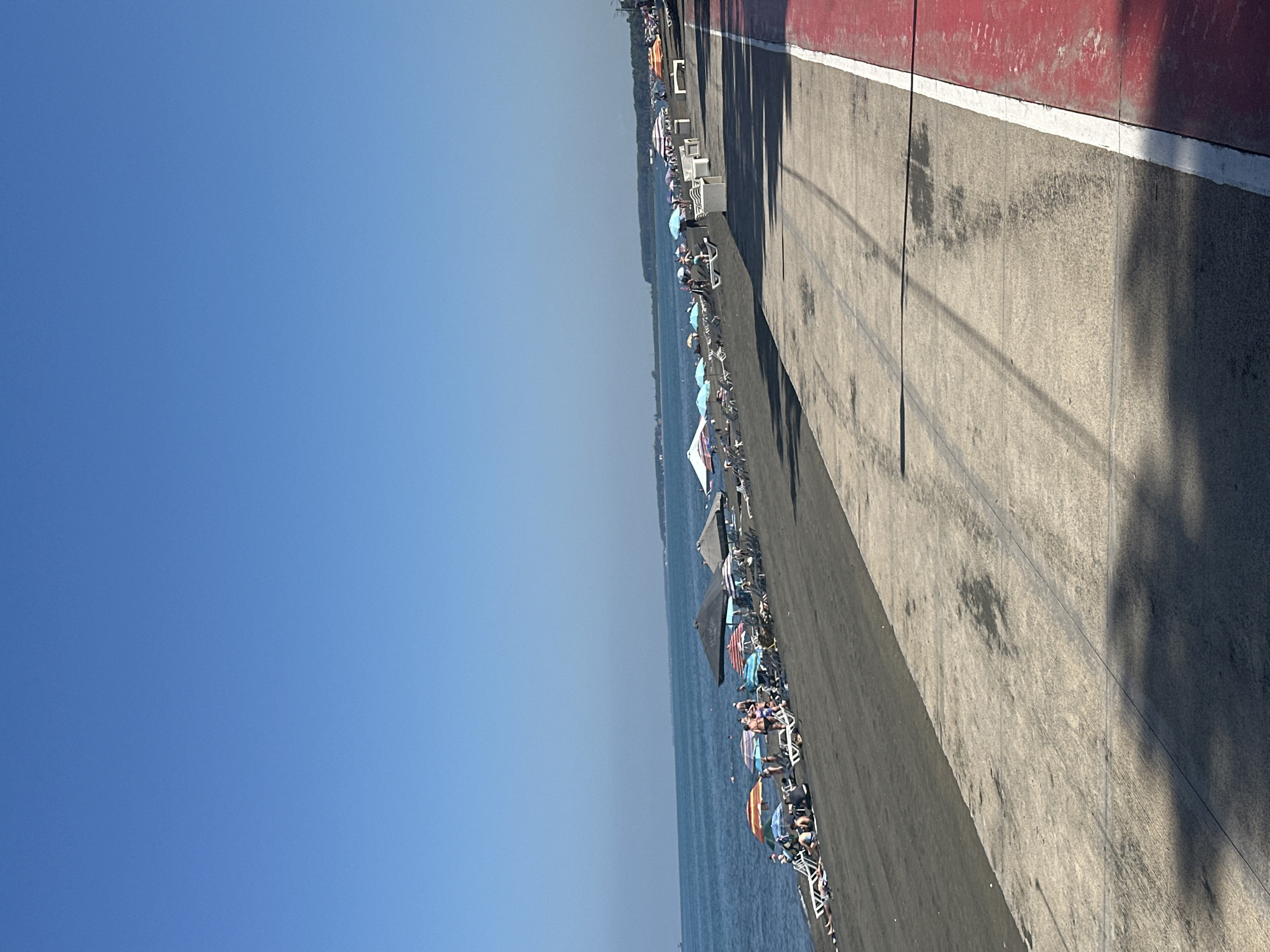 The image shows a sunny day at Ureki, Georgia, a popular Black Sea resort town known for its magnetic sand beaches. The scene is dominated by a clear blue sky and a calm, dark blue sea. The foreground features a paved walkway with a red lane separated by a white line. The walkway transitions into a dark sand beach populated with numerous beach chairs and umbrellas. Several people are visible relaxing on the beach, some under the umbrellas, while others are lying on lounge chairs. The water's edge is lined with more umbrellas and chairs, and some individuals are in the sea. In the distance, a treeline borders the coast, and the horizon is visible with a hazy outline of land. The overall impression is one of a lively and pleasant summer day at the beach.