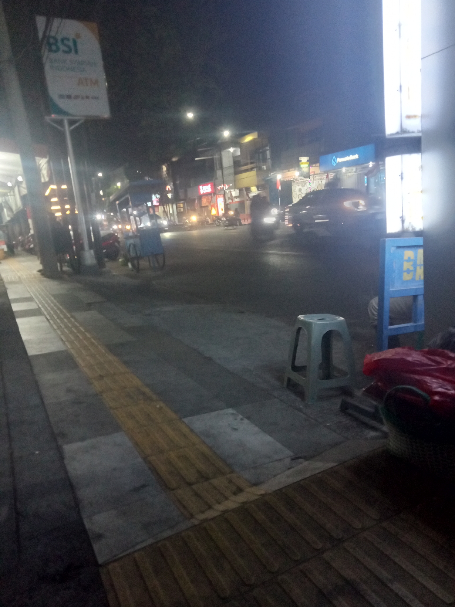 The image depicts a nighttime street scene in Probolinggo, Indonesia, likely near a BSI bank branch. A prominent BSI ATM sign is visible on a pole to the left. The street is paved with concrete and features a textured yellow tactile paving for the visually impaired along the sidewalk. Various vehicles, including cars and motorcycles, are present on the road, their headlights illuminating the scene. In the foreground, a light grey plastic stool sits on the sidewalk, next to a blue structure with yellow lettering that might be part of a kiosk or advertisement. A red plastic bag and a woven basket are also visible on the sidewalk. Buildings line the street, with illuminated signs and windows suggesting commercial activity. The overall atmosphere is that of a typical urban environment at night, with moderate street lighting.
