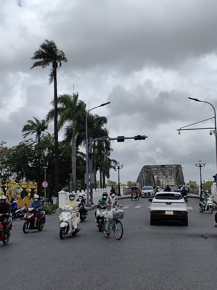 The image depicts an urban street scene on an overcast day, located in Thuận Hóa Ward, Huế, Vietnam, with the Trường Tiền Bridge visible in the background. The sky is uniformly gray with dense cloud cover.
In the foreground, an asphalt road occupies the lower third of the frame, showing signs of moisture. Numerous individuals are operating various vehicles. On the left, several motorbikes and scooters are present: a dark red scooter with a rider wearing a white helmet, a maroon scooter with a rider in a light blue jacket and white helmet, and a white scooter with a rider in a light yellow top and white helmet. Most riders are also wearing white or black face masks. A light blue bicycle with a front basket is visible in the mid-ground, ridden by a person in a patterned top, white helmet, and face mask, alongside a scooter.
In the mid-ground, a white VinFast VF 8 SUV is centrally positioned, traveling away from the viewer, displaying the license plate "75A-065.73". Traffic lights, currently unlit, are suspended over the road from a dark pole structure on the left. Multiple streetlights with dark, rectangular luminaires extend overhead on both sides of the road. A white concrete railing with ornate balusters lines the left side of the street, behind which numerous tall palm trees and dense green foliage are visible. A yellow and orange decorative structure with lantern-like elements is partially seen further left. A red "no entry" sign is also partially visible on the left. On the right, security cameras are mounted on a pole.
In the background, the metallic, arched truss structure of the Trường Tiền Bridge spans across the frame. Multiple vehicles, including cars, motorbikes, and a three-wheeled cyclo, are transiting the bridge. The bridge is also flanked by decorative streetlights similar in style to those on the foreground street.