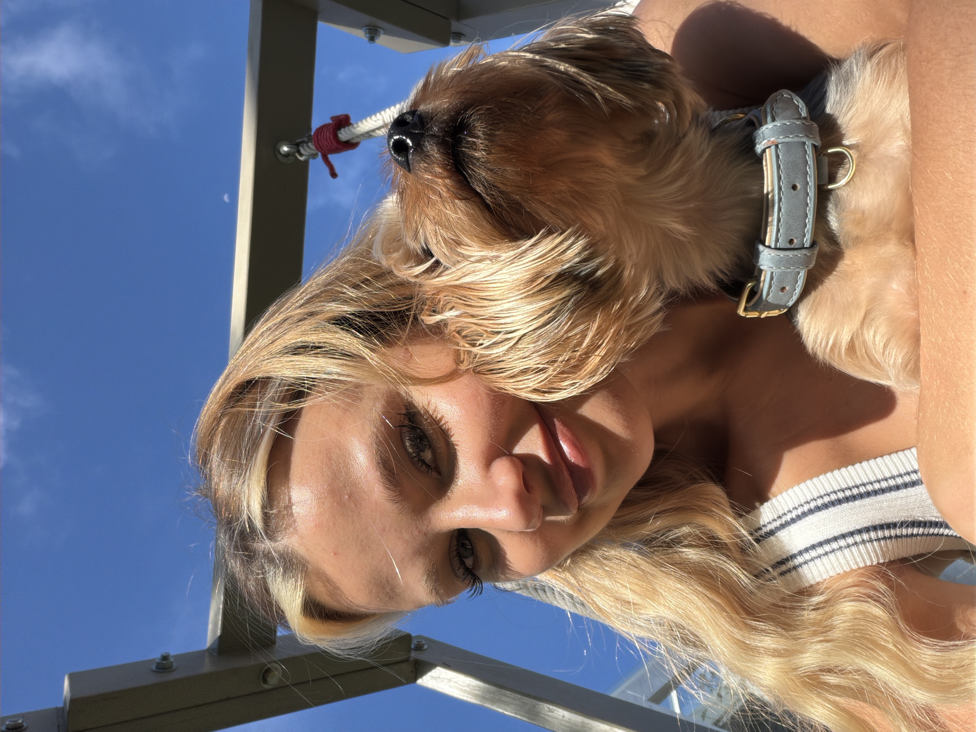 A young woman with blonde hair and a striped shirt is taking a selfie outdoors with her dog. The dog, a small brown Yorkshire Terrier, is wearing a light blue collar and is positioned close to the woman's face, looking upwards. The background features a bright blue sky with some clouds and a glimpse of a structure that appears to be part of an outdoor play area. The lighting suggests it is daytime and sunny. The image does not contain any information about the location being in Thep Krasatti, Thailand.