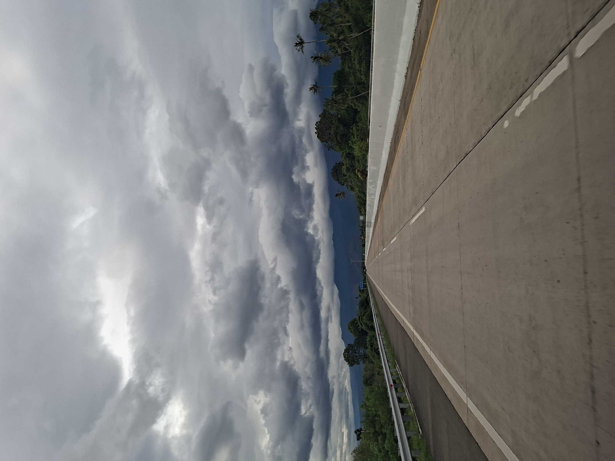 The image depicts a road in Baksukon, Indonesia, with a view of dense, ominous clouds overhead. The road itself is a grey concrete highway with a white line marking the edge. A barrier runs along the side of the road. Beyond the barrier is lush green vegetation, including trees and some palm trees. The sky is filled with dramatic, dark grey clouds, suggesting an impending storm.