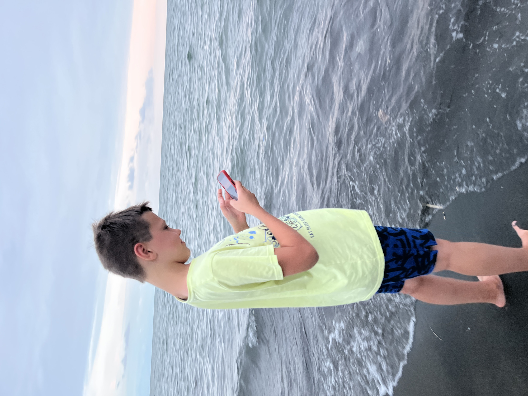 A young boy stands on a dark sand beach, facing right and looking down at a red smartphone he is holding in his hands. He is wearing a bright yellow t-shirt with a graphic print and blue patterned shorts. The ocean stretches out before him, with small waves lapping at the shore. The sky above is cloudy with hints of pink and orange, suggesting it is either dawn or dusk. The scene appears to be a tranquil setting by the sea in Ureki, Georgia.