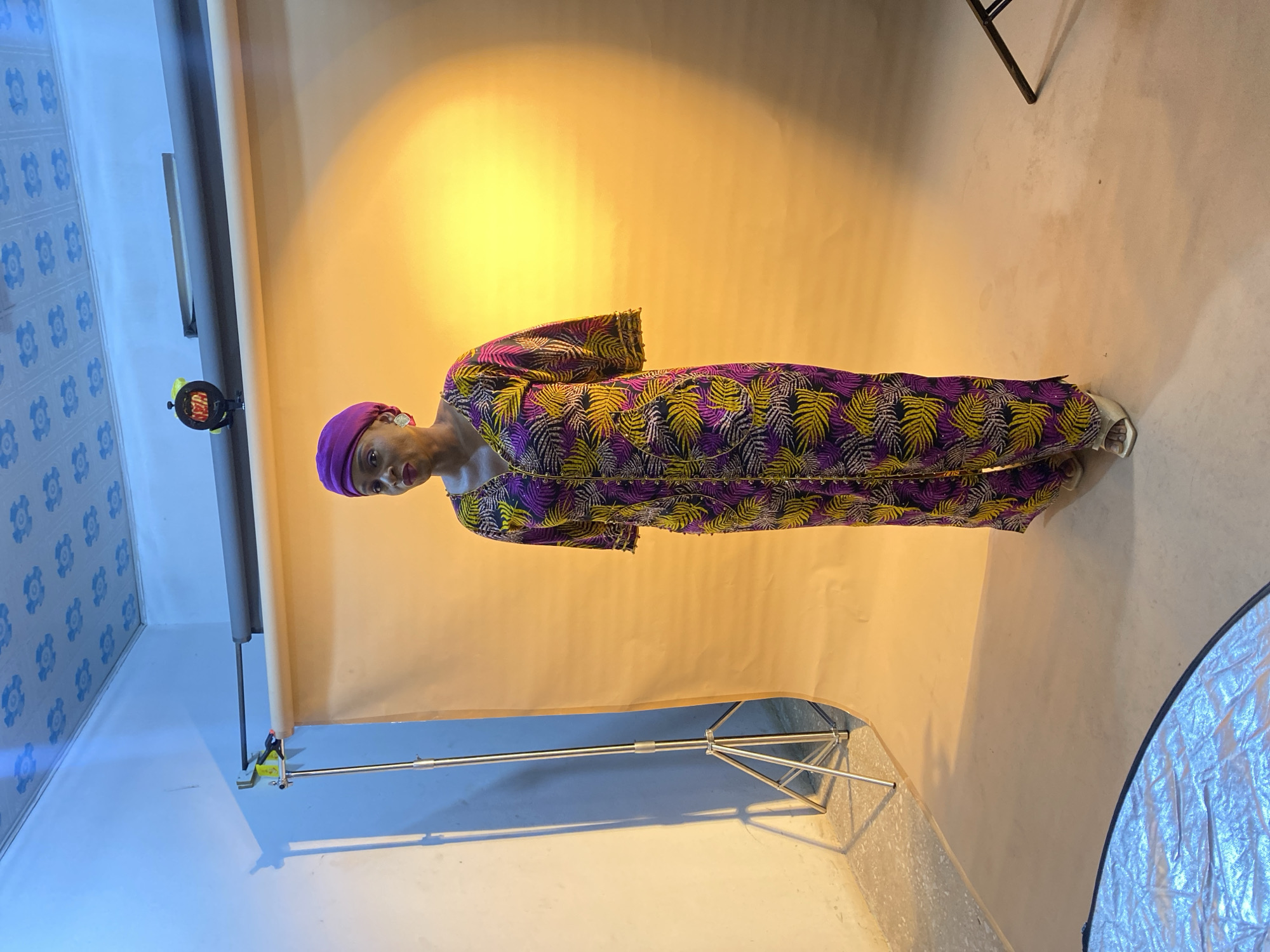 A woman wearing a purple head wrap and a long patterned dress with yellow and purple leaf-like designs is standing in front of a neutral-colored backdrop. The backdrop is supported by a metal stand. A photography reflector is visible in the bottom right corner of the frame. The room appears to be a studio or an indoor setting with blue patterned walls on one side. A digital meter or timer is attached to the backdrop stand. The location is Gura Top, Nigeria.