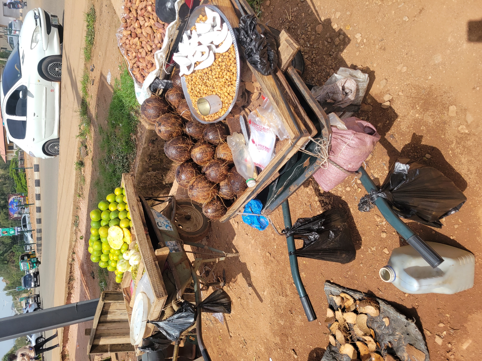 A street vendor's cart in Unknown, Unknown is pictured. The cart displays coconuts, tiger nuts, coconut flakes, dates, and limes. The vendor's cart is on a dirt area next to a street with traffic. The foreground is brown dirt.