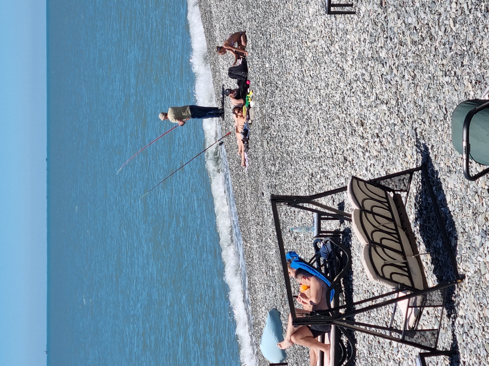 The image shows a beach scene in Sochi, Russia. The shoreline is composed of small, light-colored pebbles, transitioning into a clear blue sea. A group of people are distributed along the beach: Some are sunbathing, while one individual is fishing near the water's edge with two fishing rods. To the right, a couple is seated in a swing chair. The scene suggests a warm, sunny day with leisure activities taking place by the sea.
