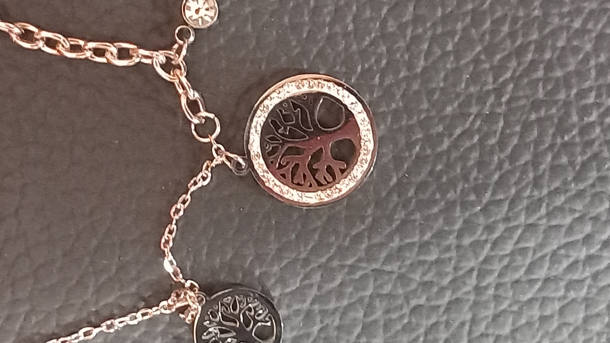 The image displays a close-up view of a rose gold colored necklace with a tree of life pendant. The necklace is lying on a dark, textured surface, possibly leather. The tree of life pendant is circular and features intricate branches within its design. The border of the pendant is adorned with small, sparkling stones, likely crystals or diamonds. A smaller, darker tree of life charm is visible on the left side of the frame, attached to the same chain. Another small, circular charm with a faceted design is present to the right of the main pendant. The lighting is dim, highlighting the metallic sheen of the jewelry. There is no indication of location within the city of Kos, Greece, nor are there any people or actions depicted.