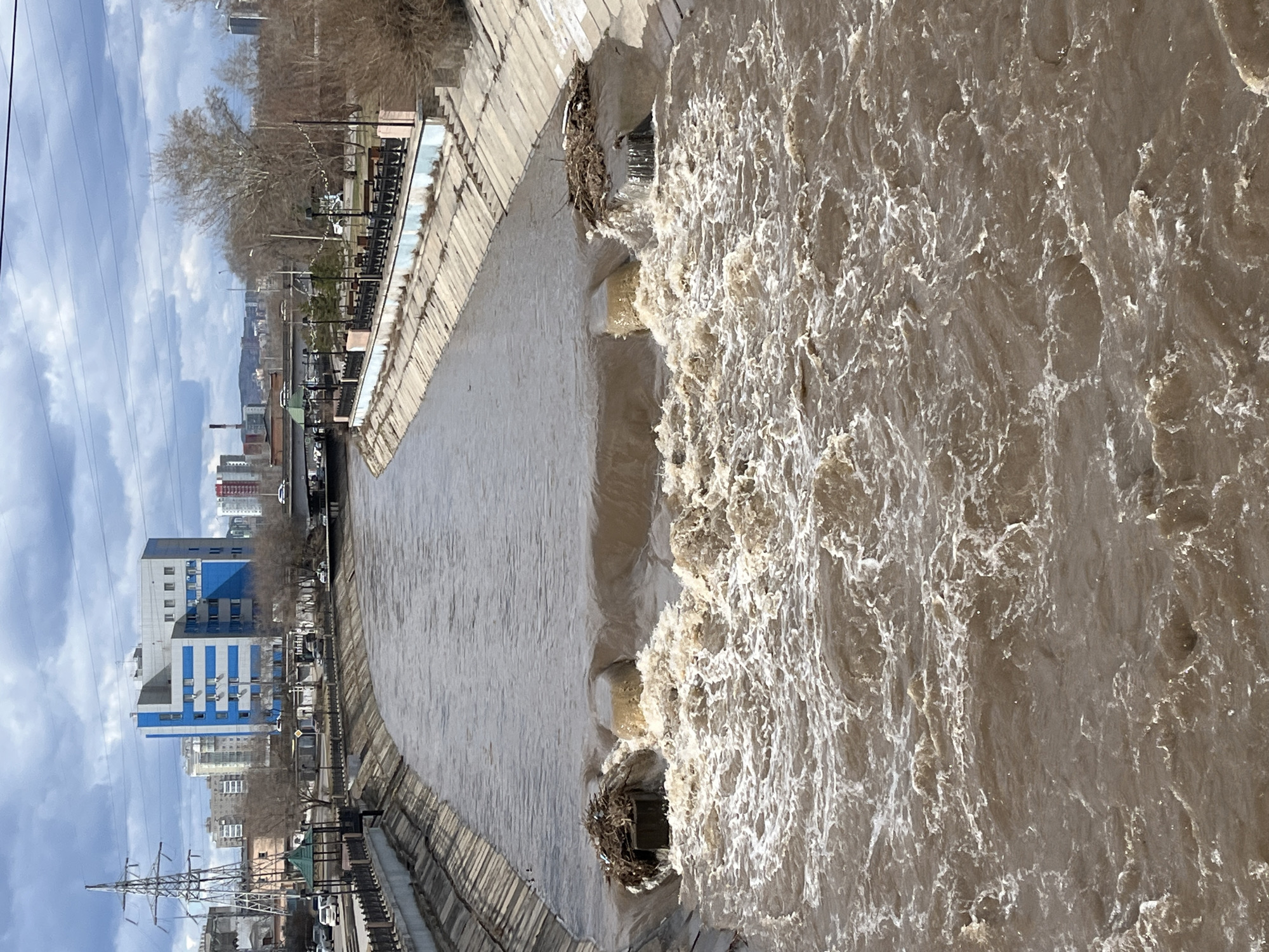 The image depicts a wide, turbulent river flowing through an urban environment in Krasnoyarsk, Russia. The river's water is a murky brown, indicating a high sediment load, and shows significant agitation with white foam and strong currents, particularly on the right side of the frame. This suggests a period of high water flow, possibly due to spring thaw or recent heavy rainfall.
The river is bordered by concrete embankments on both sides. Along the left bank, a paved area, likely a street or promenade, is visible, lined with bare deciduous trees and streetlights. Beyond this, an urban cityscape unfolds, featuring various buildings, including a distinctive multi-story structure with blue and white facades. Other buildings, some industrial and some residential, extend into the hazy distance, with hills or low mountains forming the backdrop.
Overhead, power lines traverse the sky, and a large metal power pylon is visible in the lower left portion of the image. The sky is partly cloudy, with patches of blue visible amidst the white and grey clouds, indicating a daytime scene. Debris, such as branches and twigs, is caught against submerged structures near the riverbank, underscoring the strong current. The scene captures the powerful dynamics of a river interacting with its urban surroundings during a high-water event. No discernible text is visible within the image.