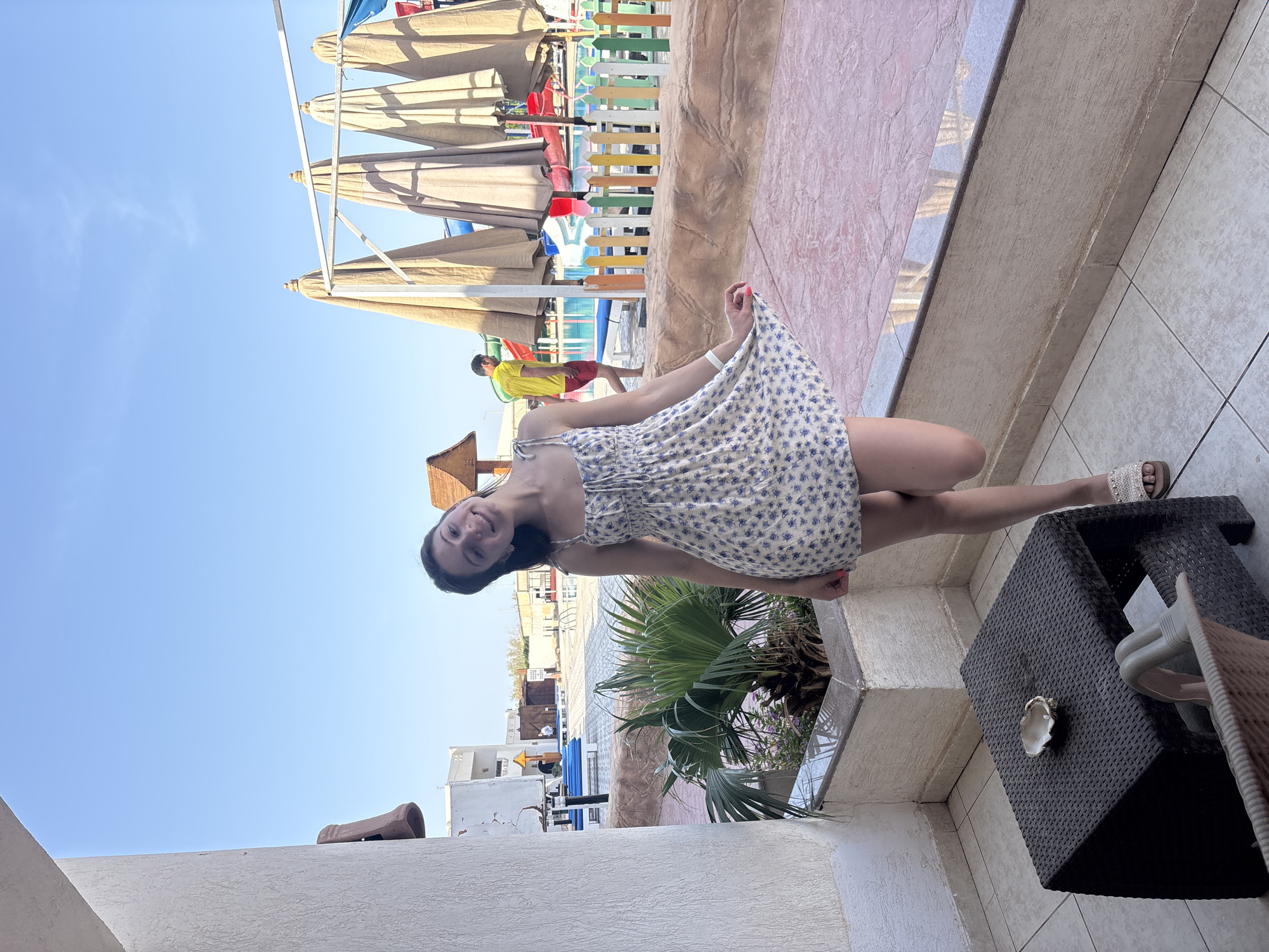 A woman in a light-colored dress with a floral pattern is standing on a tiled balcony with a dark woven table. She is holding up the hem of her dress, possibly in a playful gesture. In the background, there's a pool area with sun umbrellas, colorful fences, and a person in a yellow shirt walking near the pool. A potted palm tree sits near the edge of the balcony. The scene appears to be at a resort or hotel.