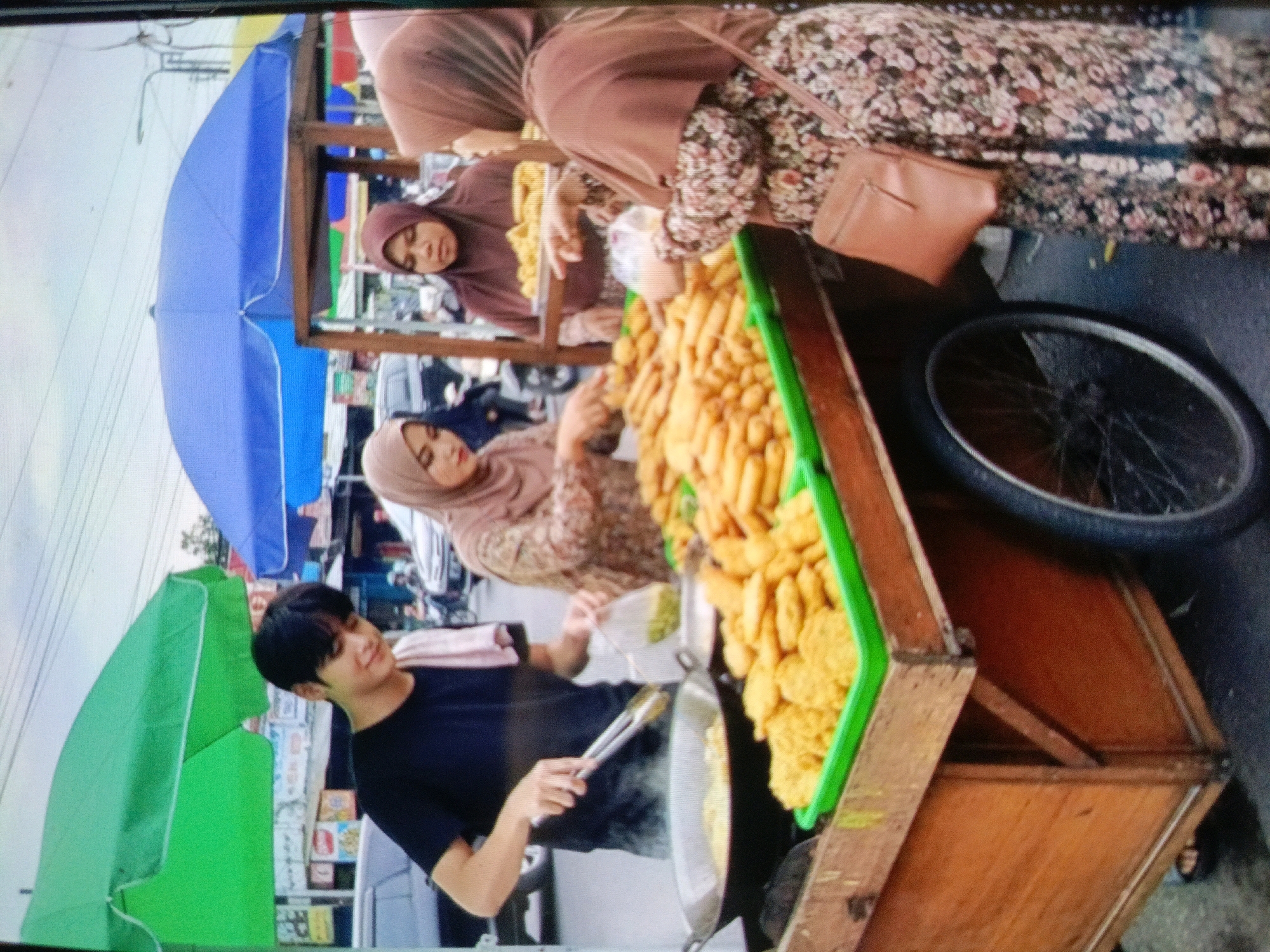 In Jenu, Indonesia, two women, likely vendors, are at a street food stall. One woman, wearing a hijab and a floral dress, is standing behind a cart filled with fried food. She holds a small purse. The other woman, also wearing a hijab, is partially obscured by the first and appears to be interacting with her. A young man in a black t-shirt stands in front of the cart, holding tongs over a pan. The cart itself is made of wood and has a green container filled with fried items. A blue umbrella is positioned to the left of the scene, and a green umbrella is on the far left. In the background, a bicycle wheel is visible, and the scene suggests a bustling street environment.