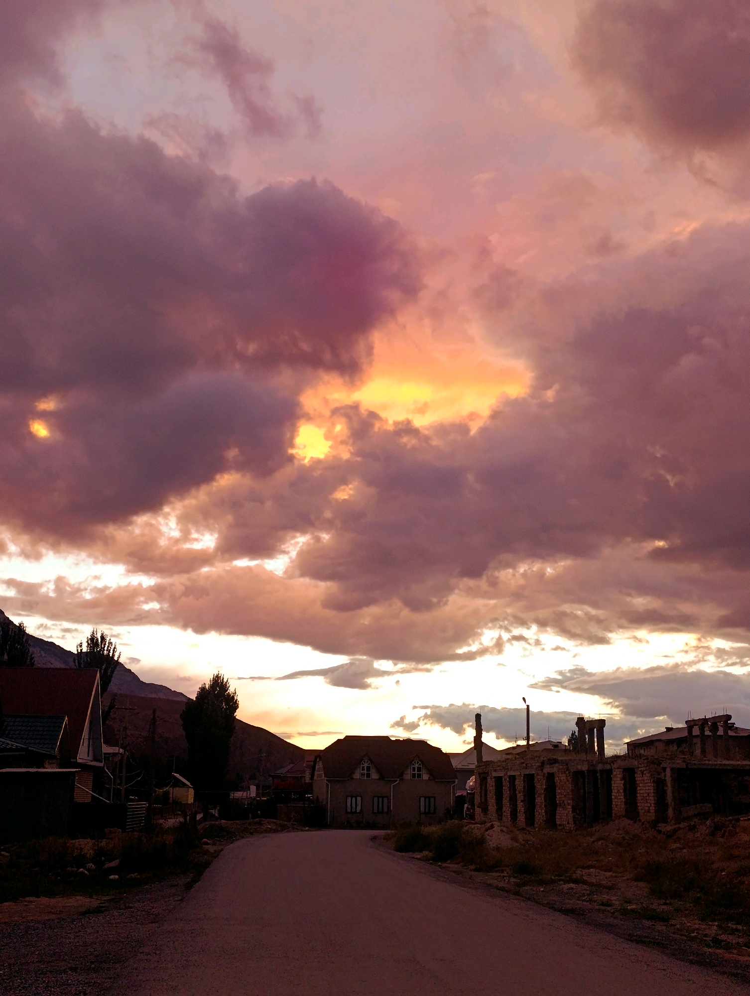 The image captures a rural village scene in Naryn City, Kyrgyzstan, at dusk. The sky is filled with dramatic, heavy clouds in shades of purple and pink, illuminated by the warm, orange glow of the setting sun breaking through. In the foreground, a paved road curves into the distance, bordered by sparse vegetation and gravel. To the left, several houses are visible, one with a reddish-brown roof. Further along the road, to the right, stands an unfinished brick structure with exposed columns and a damaged roofline. In the middle ground, a few trees stand silhouetted against the colorful sky. The mountainous terrain of Kyrgyzstan is visible in the background, adding to the dramatic and somewhat stark atmosphere of the scene. There are no people visible in the image.