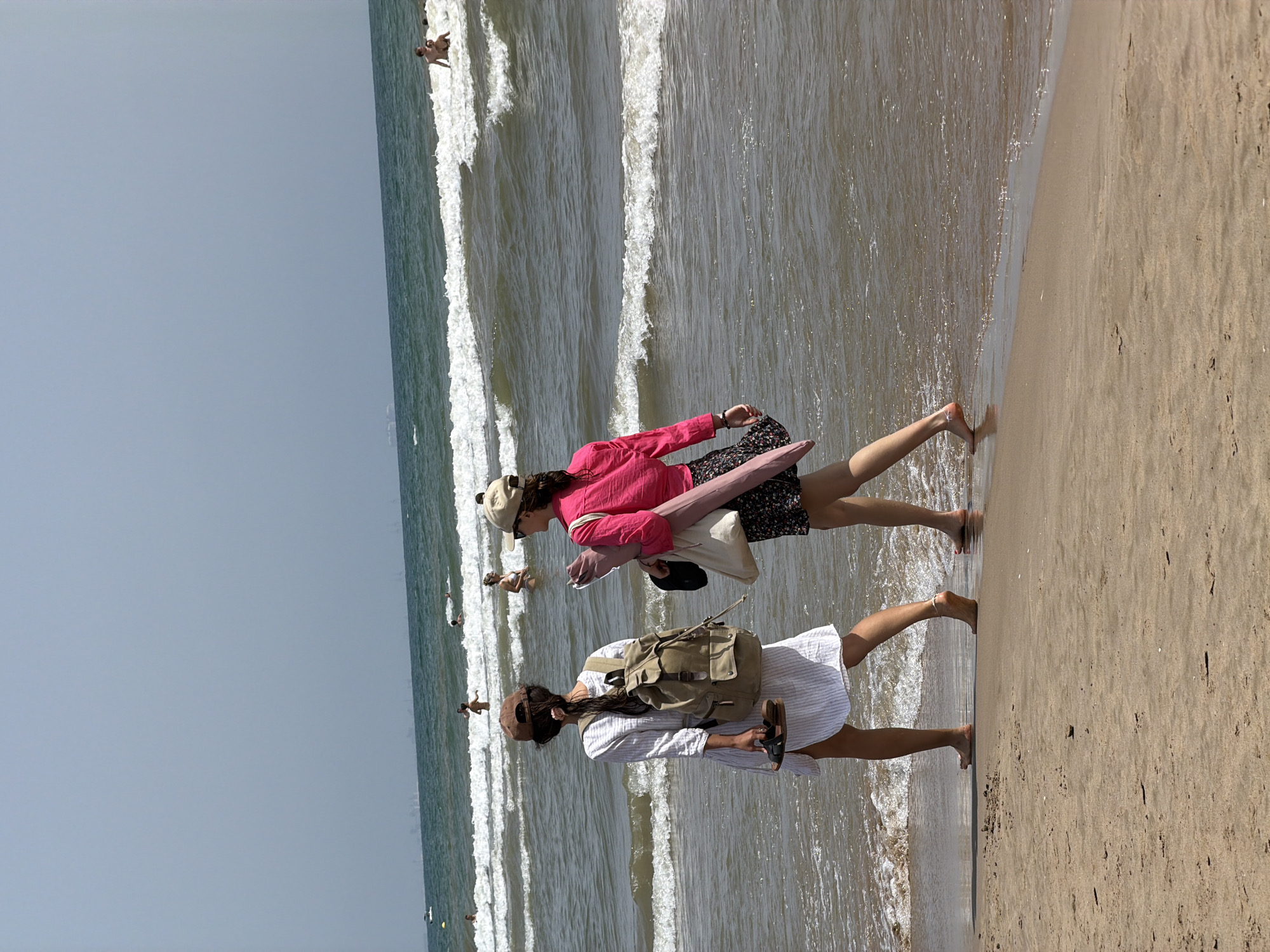 The image depicts two women walking along a sandy beach, with the ocean in the background. The woman in the foreground is wearing a white, pinstriped dress and a backpack. She is carrying sandals in her hand. The woman behind her is wearing a pink shirt and a floral skirt. She is carrying a bag and a rolled-up mat.
The scene appears to be set during the daytime, likely a warm, sunny day, as indicated by the bright lighting and the presence of people at the beach. The waves in the ocean suggest a breezy or active sea. Several other individuals can be seen in the water and on the shore in the background, some of whom appear to be swimming or wading. The sky is a pale blue, indicating clear weather. The overall atmosphere seems relaxed and leisurely, consistent with a beach outing. There is no visible text in the image.