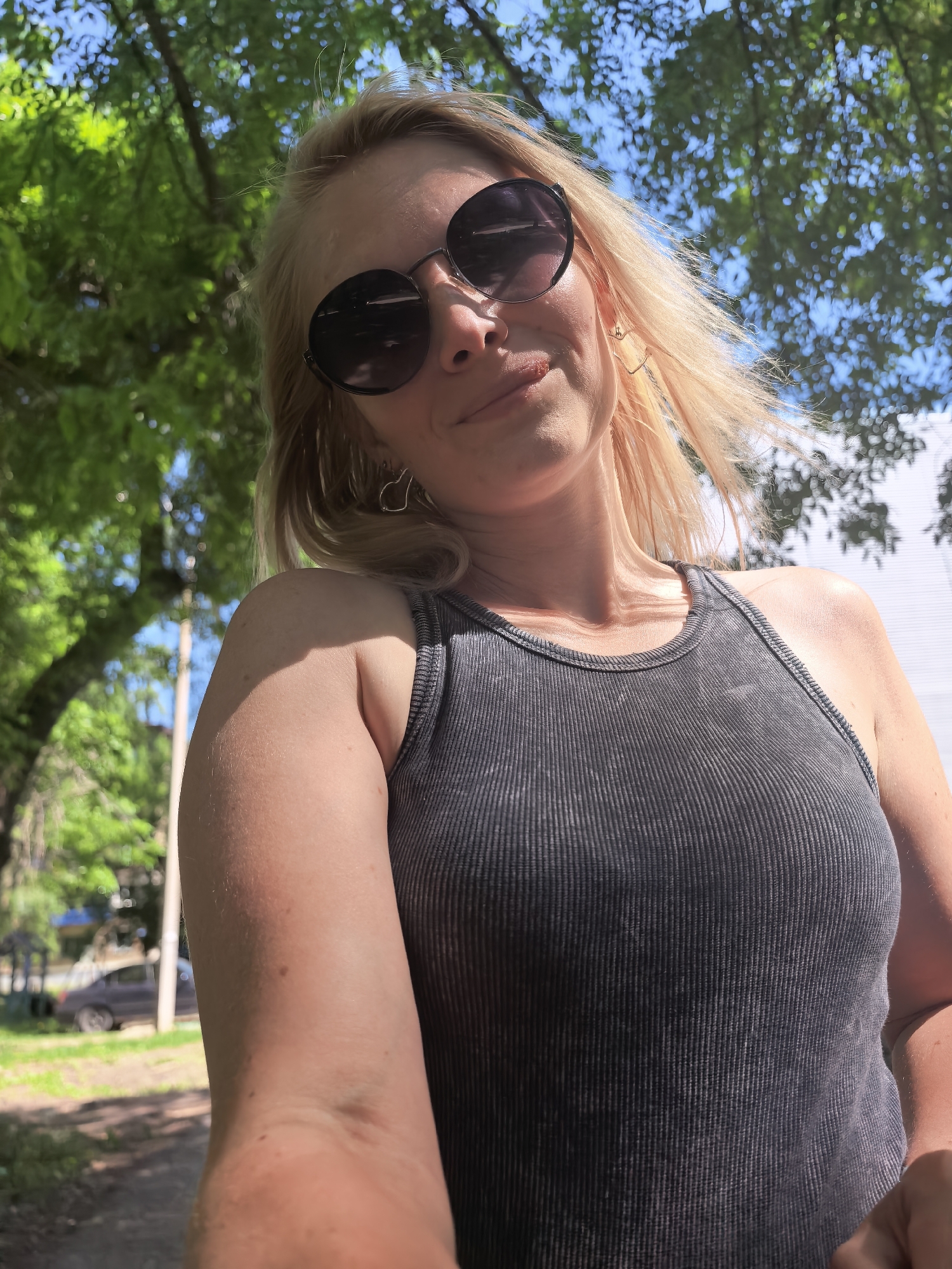 A young woman with blonde hair and sunglasses stands outside on a sunny day. She is wearing a gray ribbed tank top and heart-shaped earrings. A blurry green tree canopy is above her, with glimpses of a blue sky. In the background, on the left, is a street, part of a car, a lamp post, grass and trees and on the right, a building. The location might be the city of Sterlitamak in Russia.