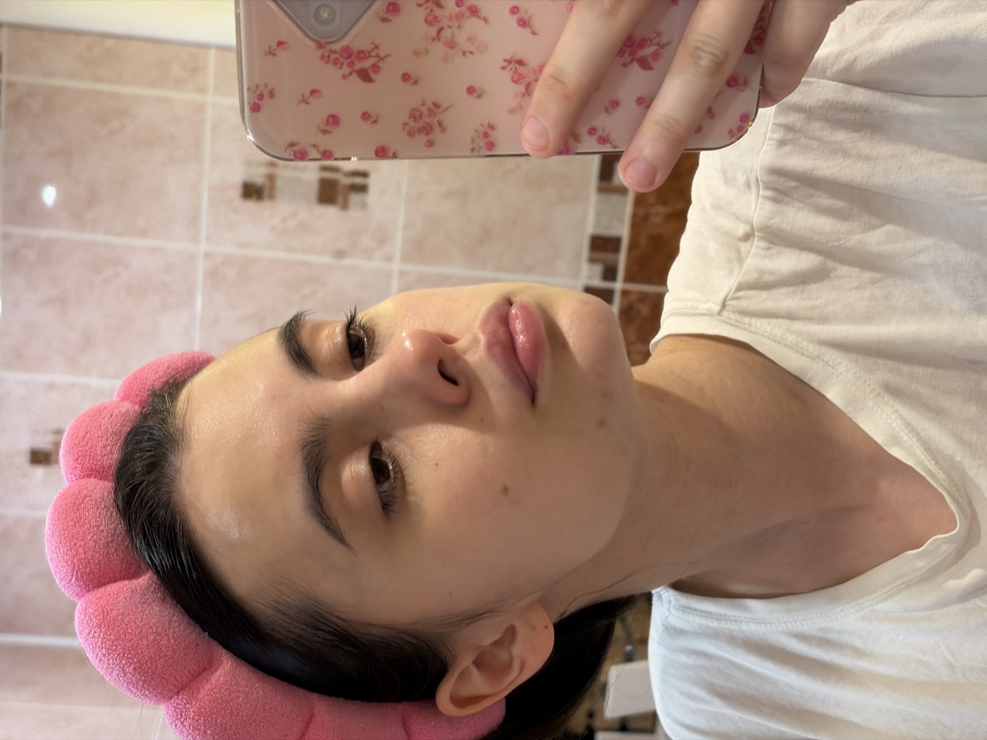 A person is holding a smartphone in selfie mode. The phone has a pink floral patterned case. The person's face is visible, with a pink headband in their hair. They are wearing a white t-shirt. The background shows a tiled wall, possibly in a bathroom. There is no indication of the location being Chaikovsky, Russia.