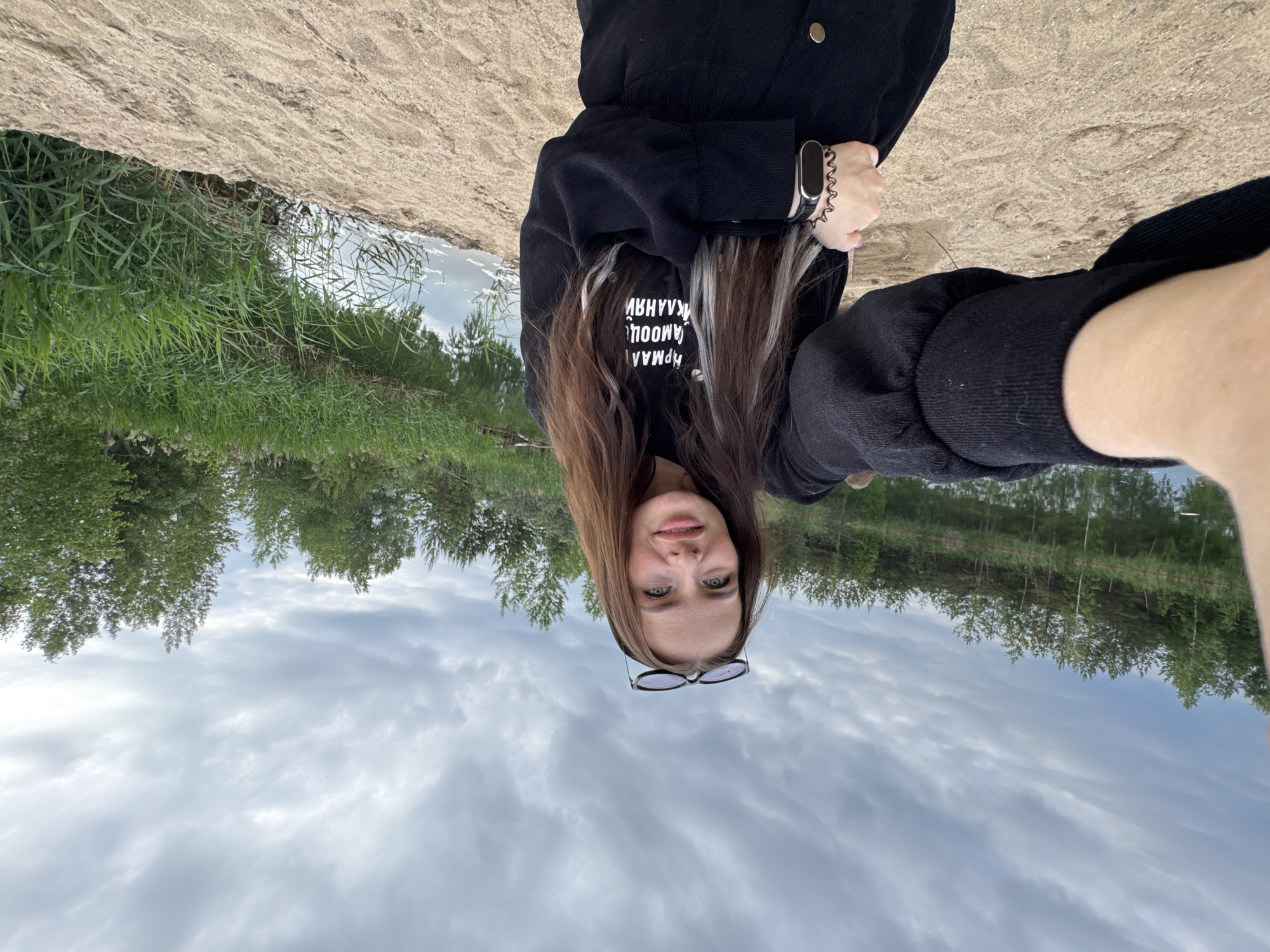 A young woman with long, brown hair and streaks of blonde, wearing a black sweatshirt, is taking a selfie by a body of water. She is wearing sunglasses on top of her head and a watch on her left wrist. The background consists of a sandy shore, dense green foliage, trees, and a cloudy sky. The location is likely a natural area or park on the outskirts of Novyi, Russia, given the natural landscape.