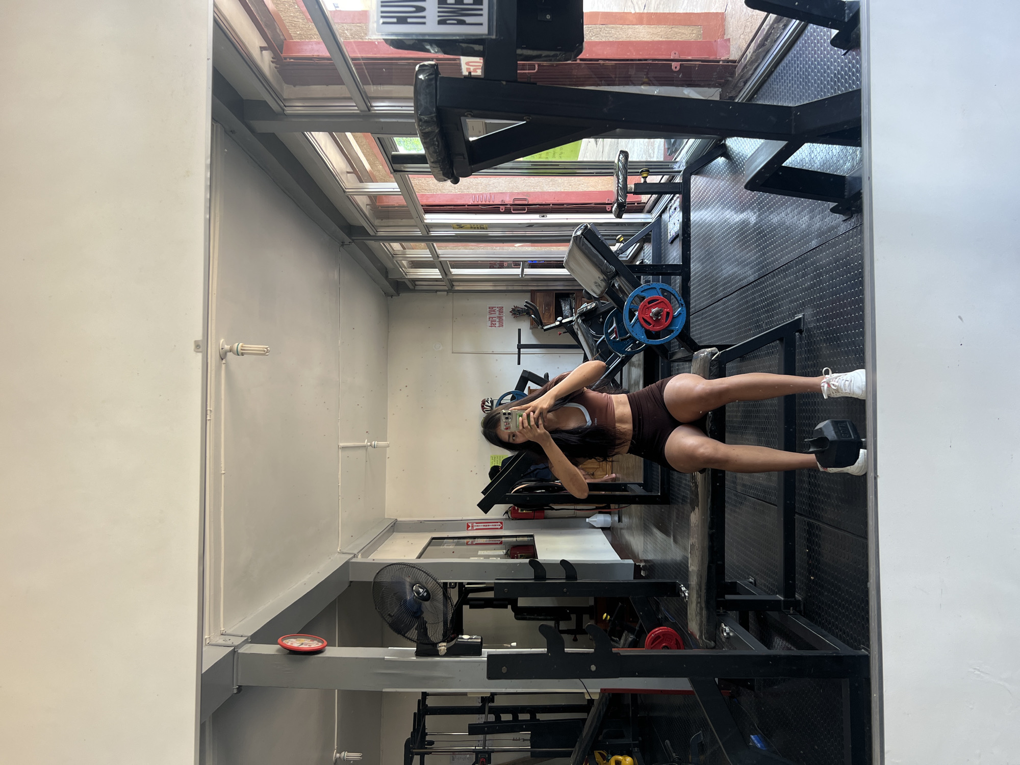 In the city of Cuyo, Philippines, a young woman is captured in a full-length mirror selfie within a gym. She is wearing a brown sports bra and brown shorts, with white sneakers. The gym appears to be equipped with various weightlifting machines and benches. A black oscillating fan is mounted on a gray pillar to the left, and a red-faced clock is visible on the same pillar. Large windows line the right side of the gym, revealing an outdoor view. The floor is covered in black textured matting. The woman is holding a smartphone in both hands, taking the photograph.