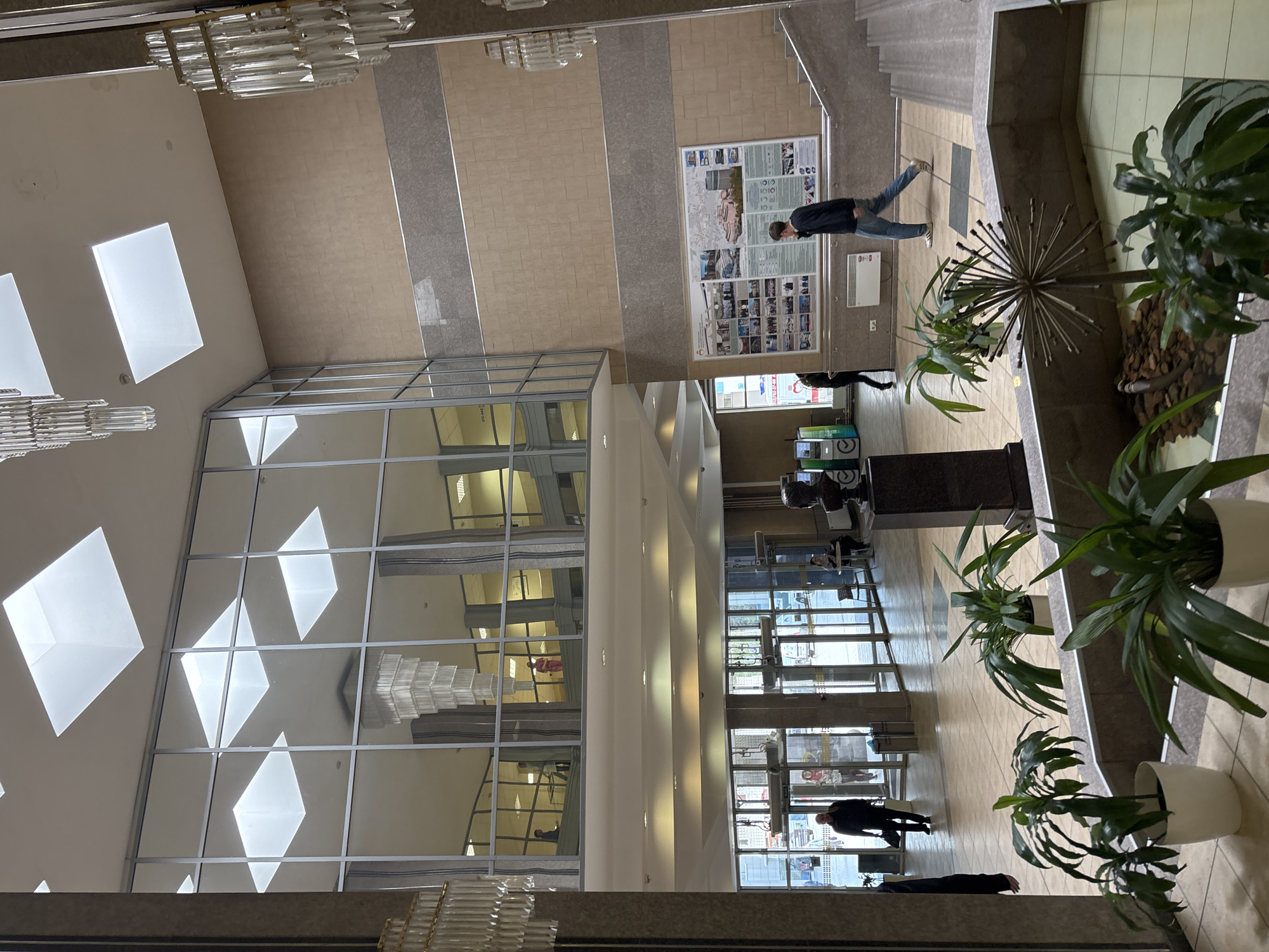 The image depicts the interior of a building, potentially a university or administrative building in Saint Petersburg, Russia. The scene is an elevated view of a multi-story atrium with a large glass wall featuring diamond-shaped skylights and chandeliers. The floor is tiled in beige and gray, with several indoor plants in white pots. People are present, including a man looking at a poster and others walking in different directions. Vending machines are visible in the background. The architecture is modern, with stairs and elevators.