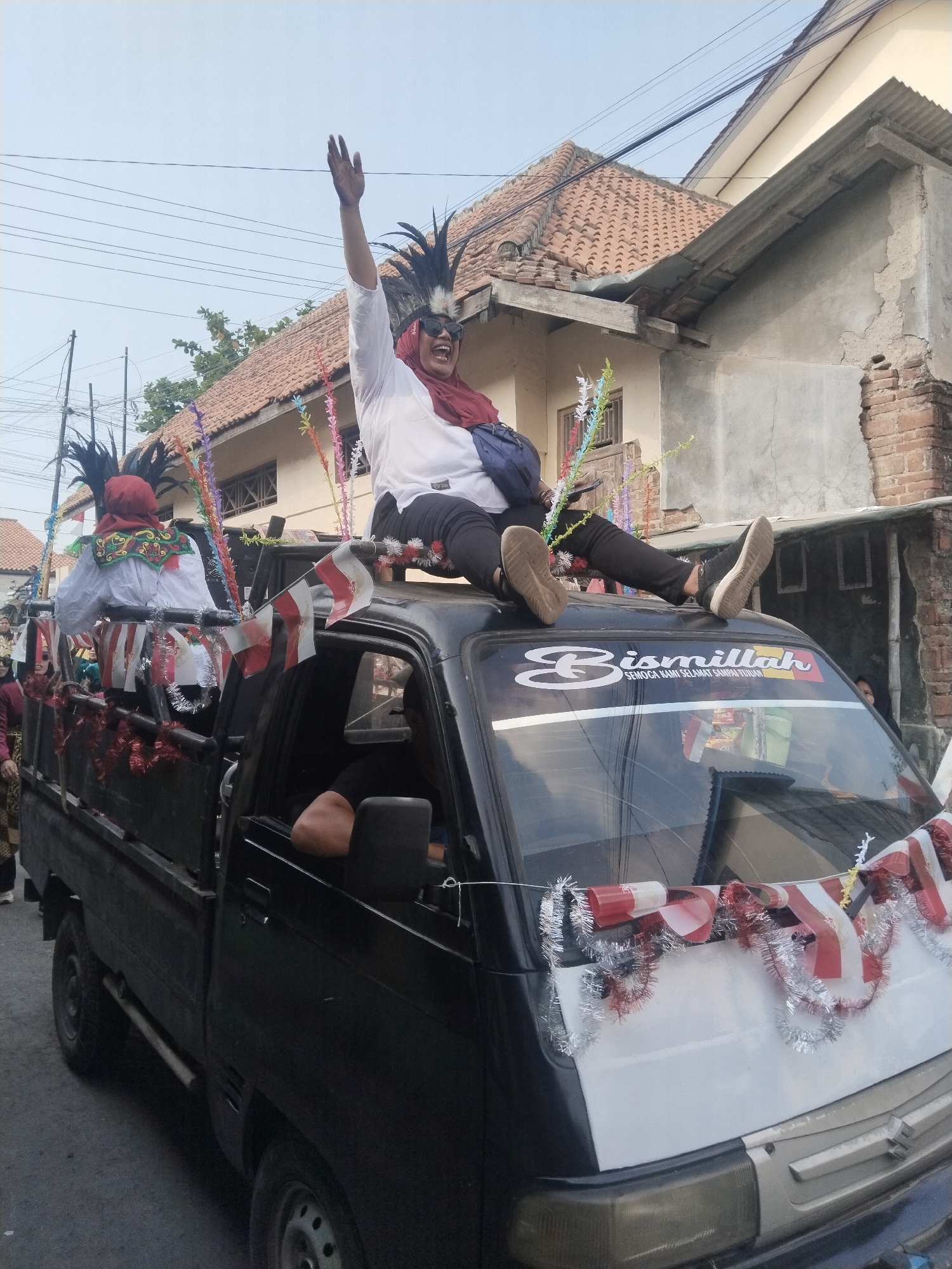 In Jembayat, Indonesia, a black pickup truck is decorated for a parade. A woman wearing a white long-sleeved shirt, a red headscarf, sunglasses, and a black headdress with feathers is sitting on the roof of the truck, waving with her right hand. She is wearing black pants and black sneakers. To her left, in the bed of the truck, another person is wearing a similar black headdress and a white shirt with a red garment around their neck. The truck is adorned with red and white Indonesian flags and tinsel. The background shows residential buildings with red-tiled roofs and overhead electrical wires. A sign on the rear windshield of the truck reads "Bismillah Semoga Kami Selamat Sampai Tujuan."