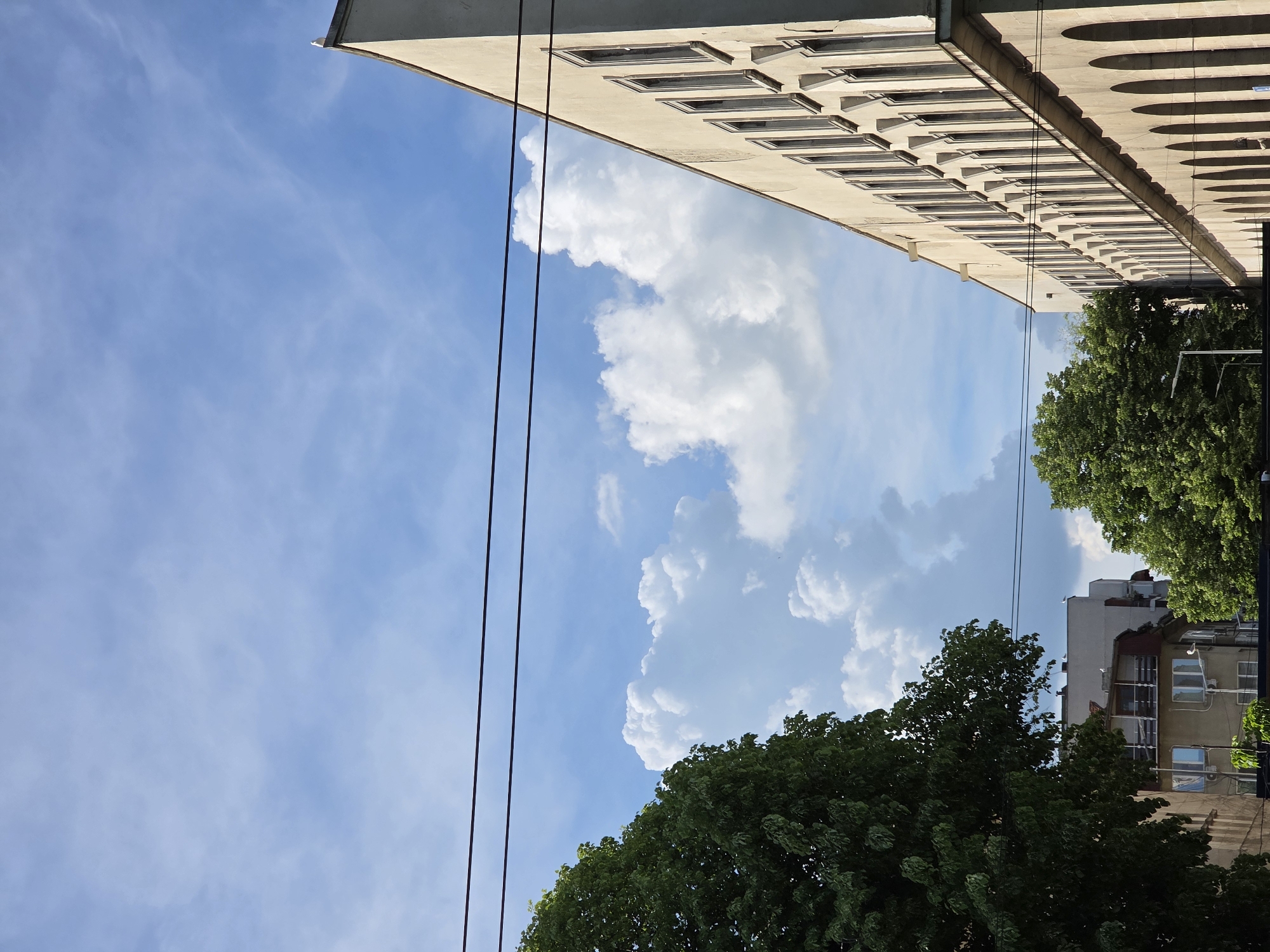 The image shows a partial view of an urban scene, angled upwards, likely taken in Varna, Bulgaria. A multi-story building with repetitive window designs and arched architectural elements occupies the upper right of the frame. Two parallel, black power lines run vertically through the left side of the image. Fluffy, white cumulus clouds fill much of the blue sky. Green foliage of trees obscures some buildings in the lower right. Other, older buildings can be seen between gaps in the tree line. The light suggests a bright, sunny day.
