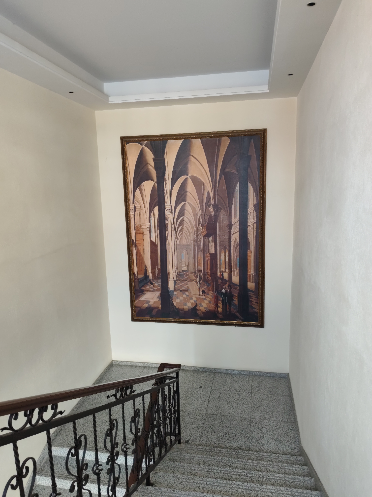 The image shows a staircase with granite treads and risers, flanked by a wrought iron railing with a dark wood handrail. The walls are painted a light cream color. A large framed painting hangs on the wall above the staircase. The painting depicts an interior architectural scene with arches and columns, rendered in a realistic style. The scene is dimly lit, with shafts of light illuminating the checkered floor and a few small figures. There are no people visible in the immediate vicinity of the staircase. The location is identified as Aihui, China.