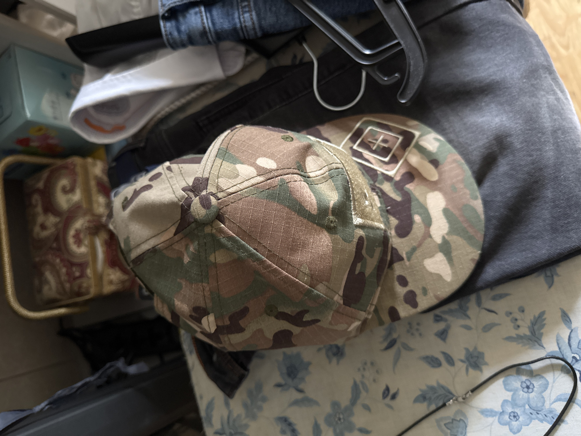 The image shows a close-up, top-down view of a camouflage baseball cap, with the front of the cap facing the viewer. The cap has a patch on the front, which appears to be a diamond shape with a cross symbol inside, in a light tan color against the camouflage fabric. The cap is lying on top of a dark gray piece of clothing, possibly denim jeans or a jacket.
To the left of the cap, partially visible, is a brown woven basket containing a patterned fabric with paisley-like swirls in red and gold. Behind the basket and cap, a light blue box with cartoonish graphics is visible, along with a white folded item. At the top of the image, a portion of dark blue denim jeans is visible. A black clothing hanger is also present, hooked over the edge of the dark gray garment. The overall setting appears to be indoors, within a closet or storage area. There is no indication of the time of day, weather, or specific emotions. No visible text is present in the image.
